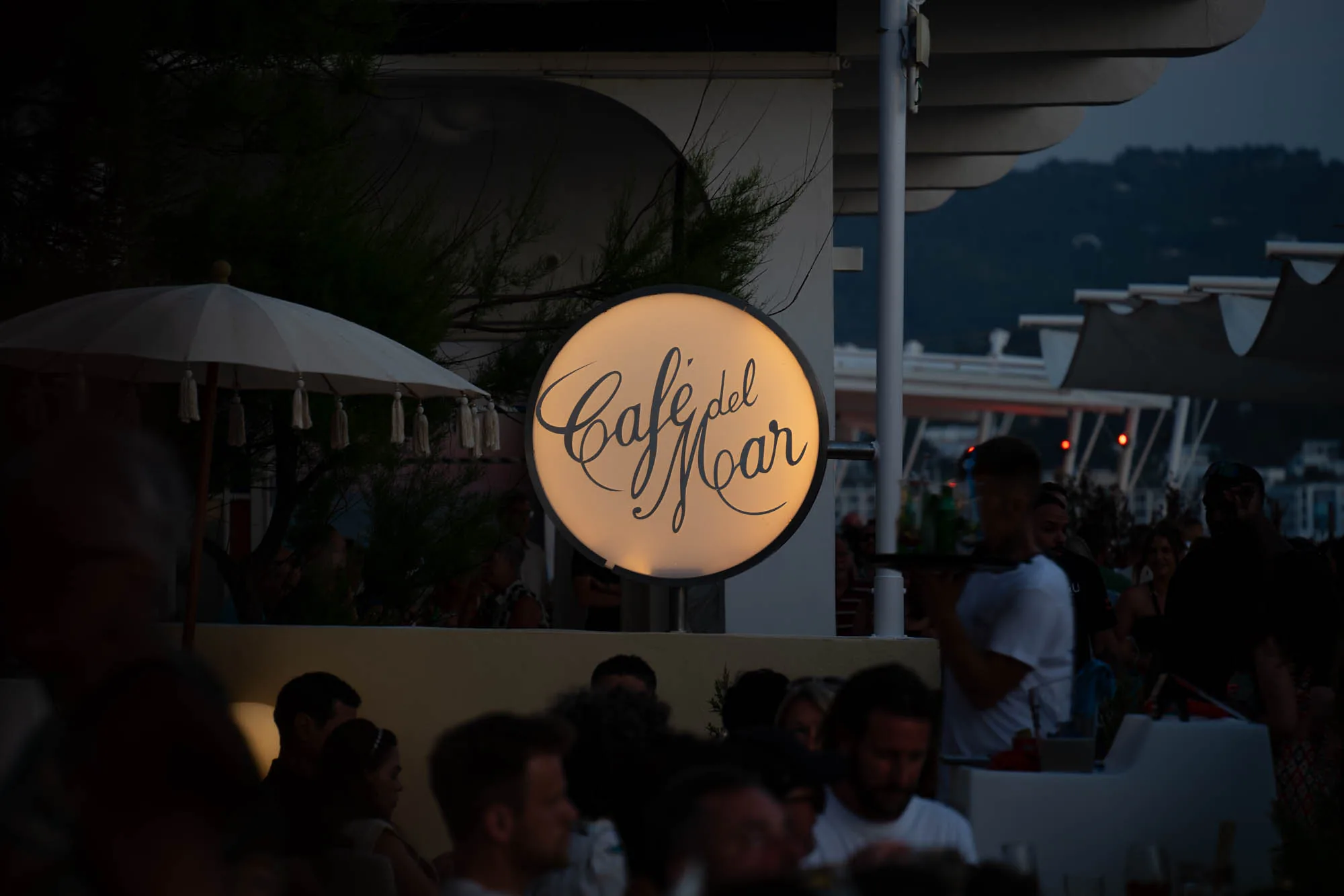 写真はスペインのイビサ島にある「Cafe del Mar」で撮影された夜の様子です。写真はやや暗めで、夕暮れ時の静かな雰囲気を醸し出しています。中央には、丸いライトサインがあり、「Cafe del Mar」の文字が黒い筆記体で書かれていて、明るく輝いています。サインの周りには、木々や白いパラソルが見えます。  写真の下部には、カフェのテラス席に座る人々の頭部が写っています。人々は会話を楽しんでいる様子で、リラックスした雰囲気が伝わってきます。背景には、海や山が見え、イビサの美しい自然を感じさせます。写真全体からは、落ち着いた雰囲気と洗練された空間が伝わり、イビサのナイトライフを垣間見ることができます。