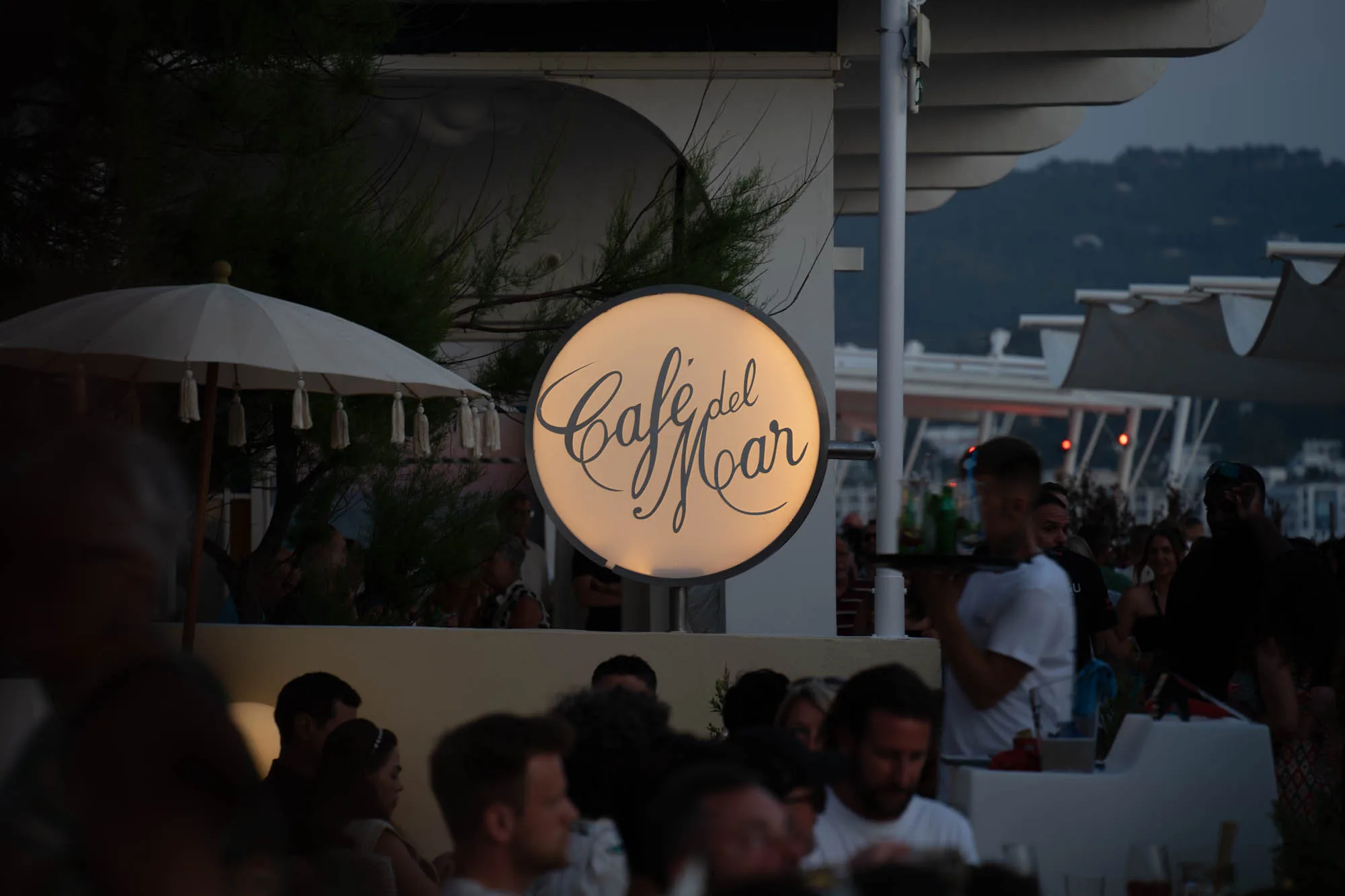 写真はスペインのイビサにある有名なカフェ「Cafe del Mar」の夜の様子を捉えています。大きく照らされた丸い看板が印象的で、店名が優雅な筆記体で書かれています。看板の周りには木々や傘が配置され、背景には人が賑わう様子が伺えます。ウェイターが飲み物を運んでいる姿も見え、リラックスした雰囲気と活気が伝わってきます。夕暮れ時の穏やかな光が全体を包み込み、落ち着いた大人の空間を演出しています。
