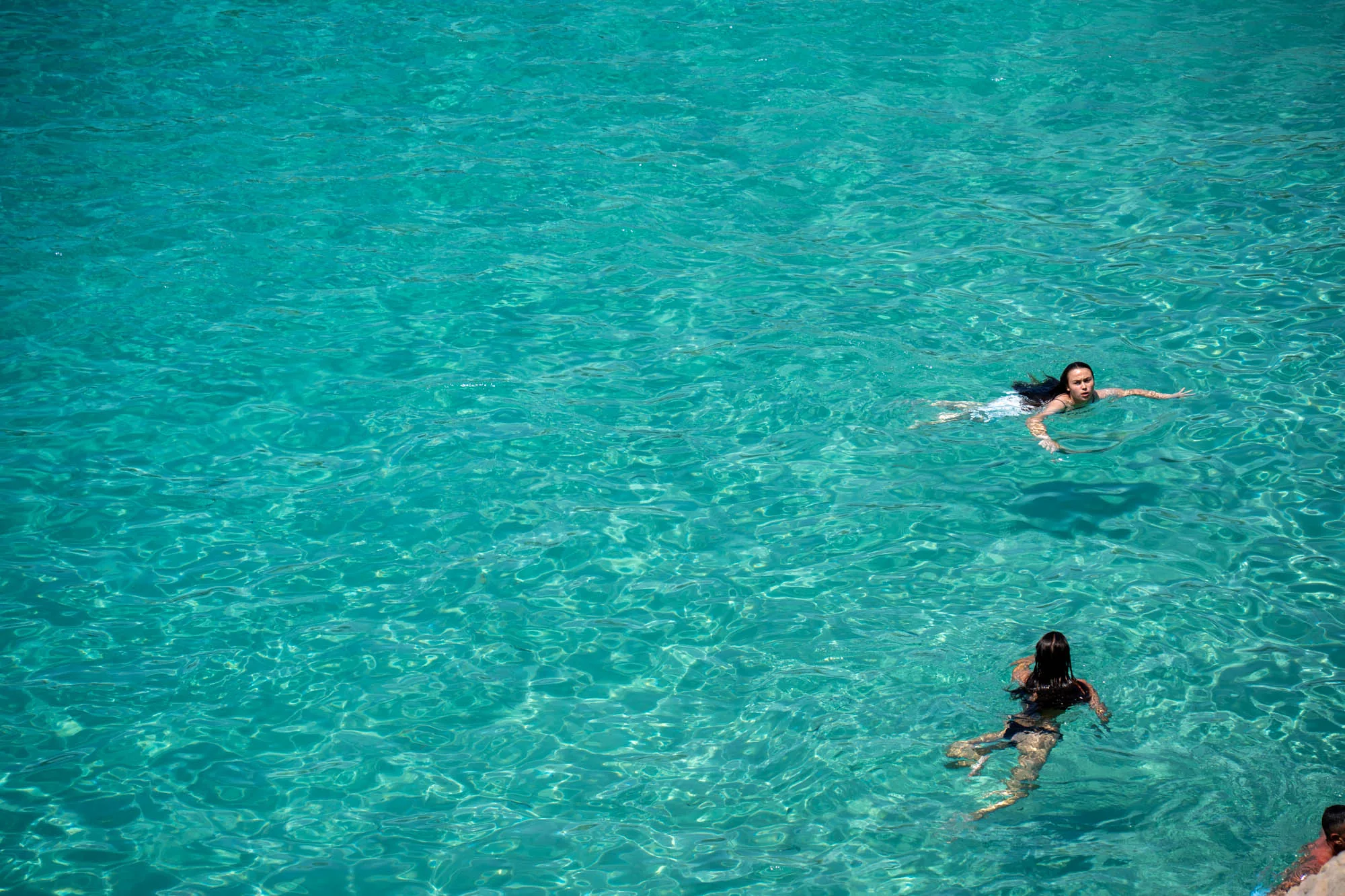 スペインのイビサで撮影されたと思われるこの写真は、鮮やかなターコイズブルーの海で泳ぐ人々を捉えています。水面は太陽光を反射してキラキラと輝き、透明度が高く、海底の模様が透けて見えます。写真の上部には、黒髪の女性がゆったりと仰向けで浮かんでいます。水着を着ているようです。写真の下部には、同じく黒髪の女性が泳いでおり、水着を着ているのが分かります。右下には男性の頭の一部が見えます。全体的に、夏のバカンスを連想させる、開放的でリラックスした雰囲気の写真です。