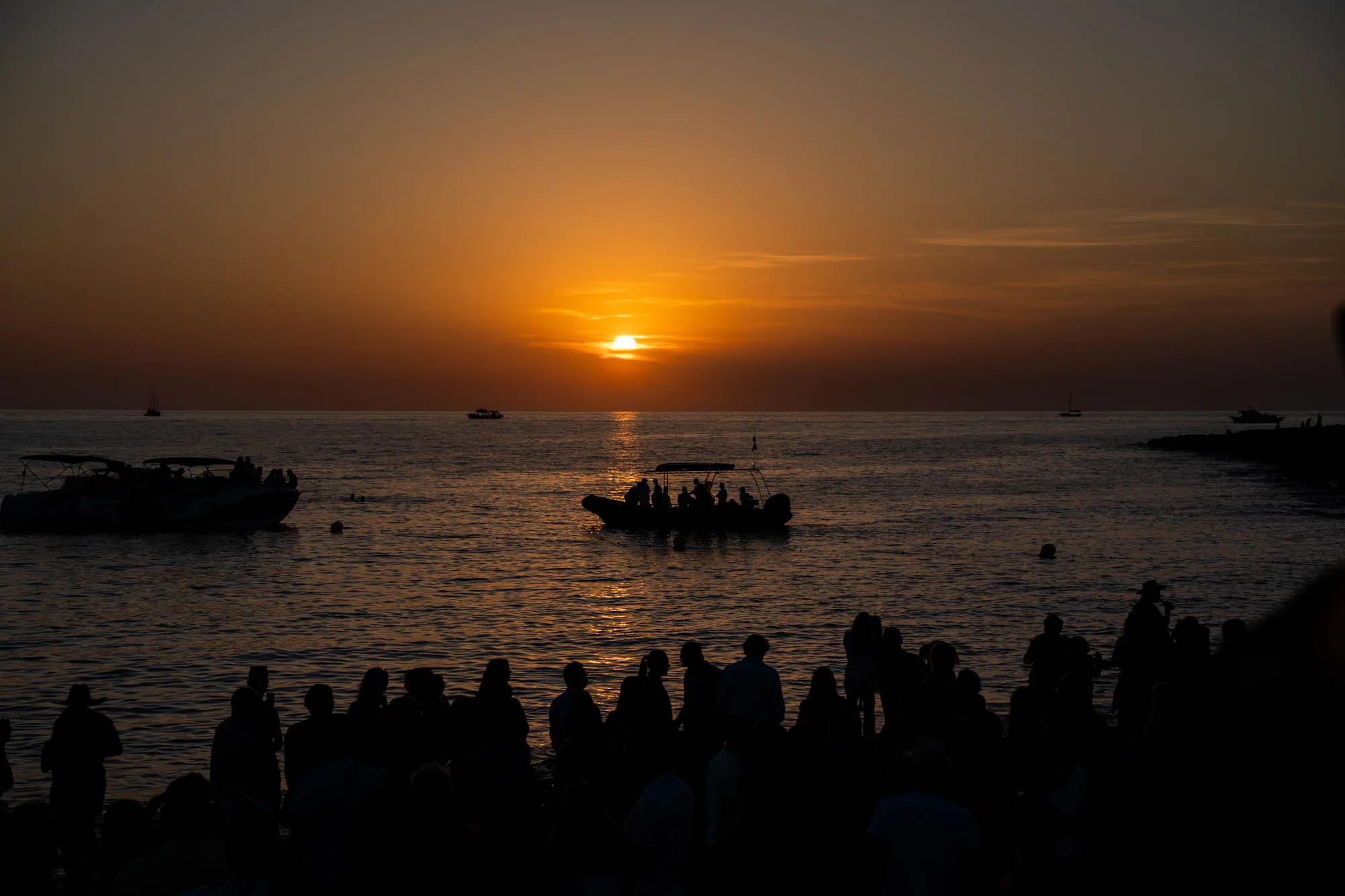 スペインのイビサで撮影された写真です。夕日が水平線に沈む瞬間の風景が捉えられています。前景には、シルエットになった人々の群れが写っており、彼らは夕日を眺めているようです。海にはいくつかのボートが浮かんでおり、そのうちの一つのボートには人が乗っているのが見えます。空はオレンジ色から暗い色へのグラデーションで、夕日の光が海面に反射して輝いています。全体的に、穏やかで美しい夕暮れ時の風景が表現されています。