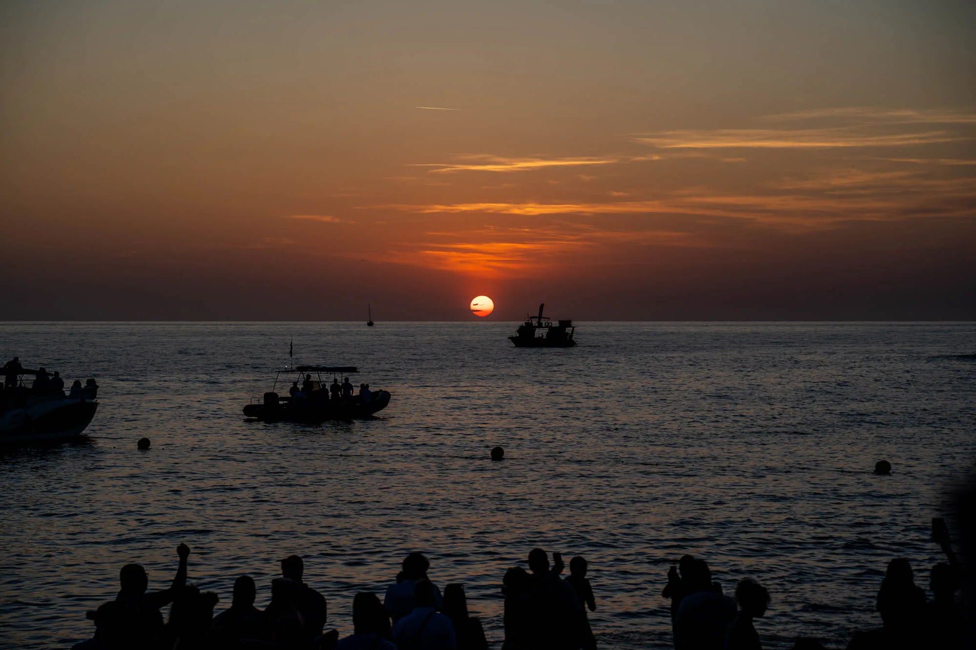 スペインのイビサ島で撮影された、夕日の写真です。太陽は水平線に沈みかけ、空はオレンジと赤のグラデーションに染まっています。海面は穏やかで、夕焼けの色を反射して輝いています。海には複数の船が浮かんでおり、シルエットになっています。手前には、海岸線に集まった人々が同様にシルエットで写っており、夕日を眺めたり、写真を撮ったりしている様子が伺えます。全体的に、穏やかで美しい夕景が捉えられています。