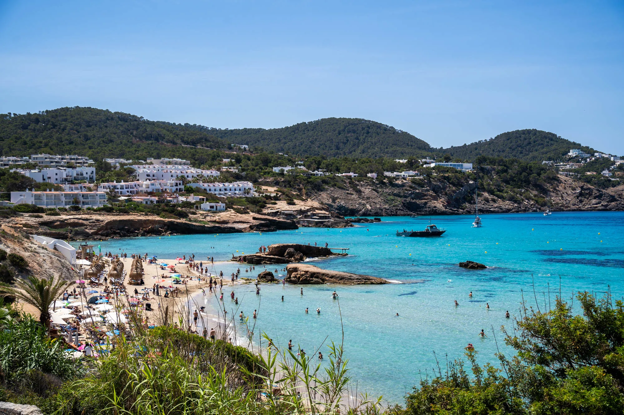 写真はスペイン、イビサ島のビーチの風景を捉えています。手前には緑豊かな植物が生い茂り、その奥には白い砂浜が広がっています。ビーチにはたくさんの人が日光浴を楽しんだり、海で泳いだりしています。海の色はターコイズブルーで、非常に透明度が高く、海底が見えるほどです。ビーチの奥には、白い建物が点在する緑豊かな丘陵地帯が広がっており、リゾート地特有の風景を作り出しています。海にはボートが浮かんでおり、穏やかな雰囲気を醸し出しています。全体的に、夏のバカンスを楽しむ人々の賑わいと、イビサ島の美しい自然が調和した、リラックスできる風景です。