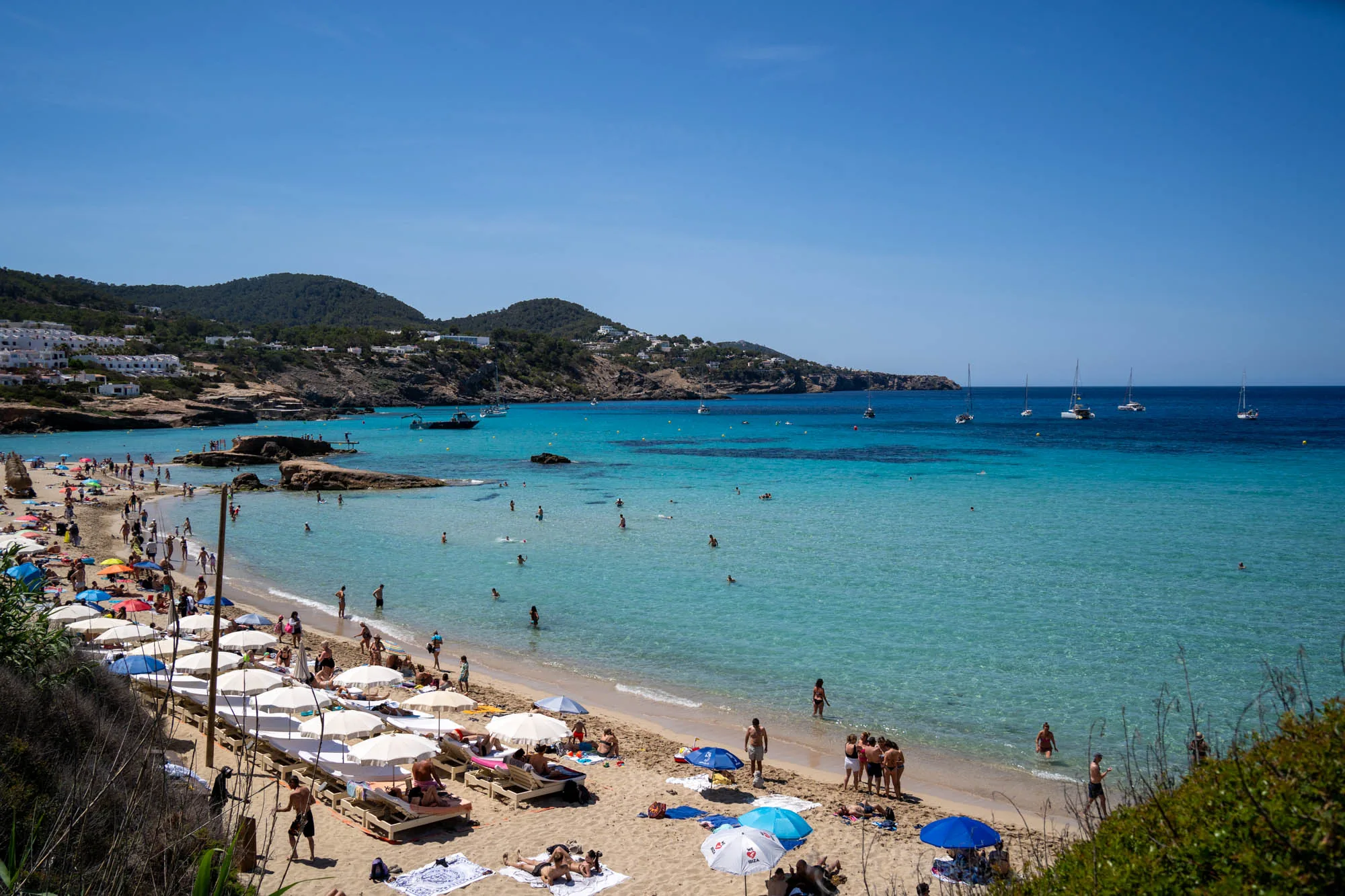 写真はスペインのイビサ島で撮影されたビーチの様子です。空は澄み切った青空で、太陽が輝いています。海はエメラルドグリーンの透明度が高く、遠くにはヨットがいくつか浮かんでいます。ビーチには多くの人が海水浴や日光浴を楽しんでおり、カラフルなパラソルが砂浜を彩っています。ビーチの奥には緑豊かな丘陵地が広がっており、白い建物が点在しています。全体的に明るく開放的な雰囲気で、リゾート地ならではの活気と穏やかさが感じられます。手前側にはビーチベッドと白いパラソルが並んでおり、リラックスした空間が広がっています。