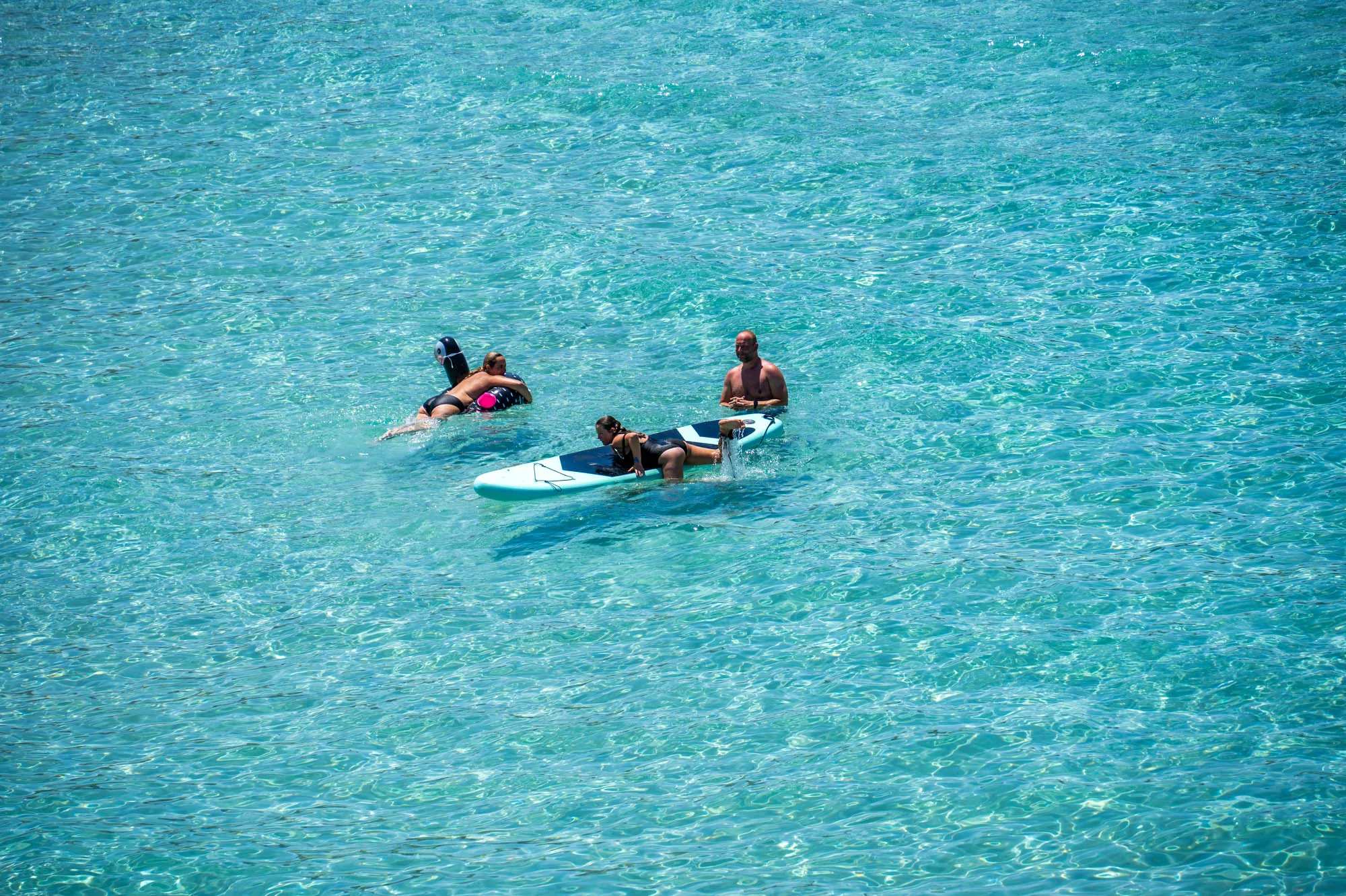スペイン、イビサ島の透き通ったターコイズブルーの海を上から捉えた写真です。画面全体に広がるのは、太陽の光を反射してきらめく水面。その穏やかな波間には、海水浴を楽しむ3人の姿が見えます。左側には、浮き輪につかまりながらリラックスした様子で浮かんでいる女性がいます。中央には、水色のサーフボードに乗って身を起こしている女性がおり、そのそばには立って海に入っている男性がいます。3人とも水着姿で、夏のバカンスを満喫している様子が伝わってきます。写真全体から、イビサ島の開放的で穏やかな雰囲気が感じられます。