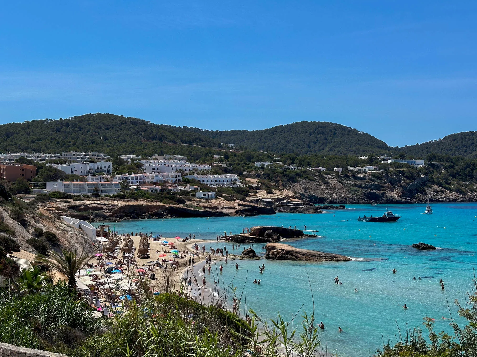 写真はスペインのイビサ島で撮影されたものです。青い空の下、ターコイズブルーの美しい海が広がっており、その海岸には多くの人々が訪れて海水浴を楽しんでいます。ビーチは砂浜で、色とりどりのパラソルが立ち並び、日光浴をしている人々や、海に入って泳いだり、水遊びをしたりしている人々が見られます。ビーチの後ろには、白い建物が並ぶ集落があり、その背後には緑豊かな山々が連なっています。海にはボートが浮かんでおり、穏やかな波が打ち寄せています。全体的に明るく、リゾート地の賑わいと活気が感じられる写真です。