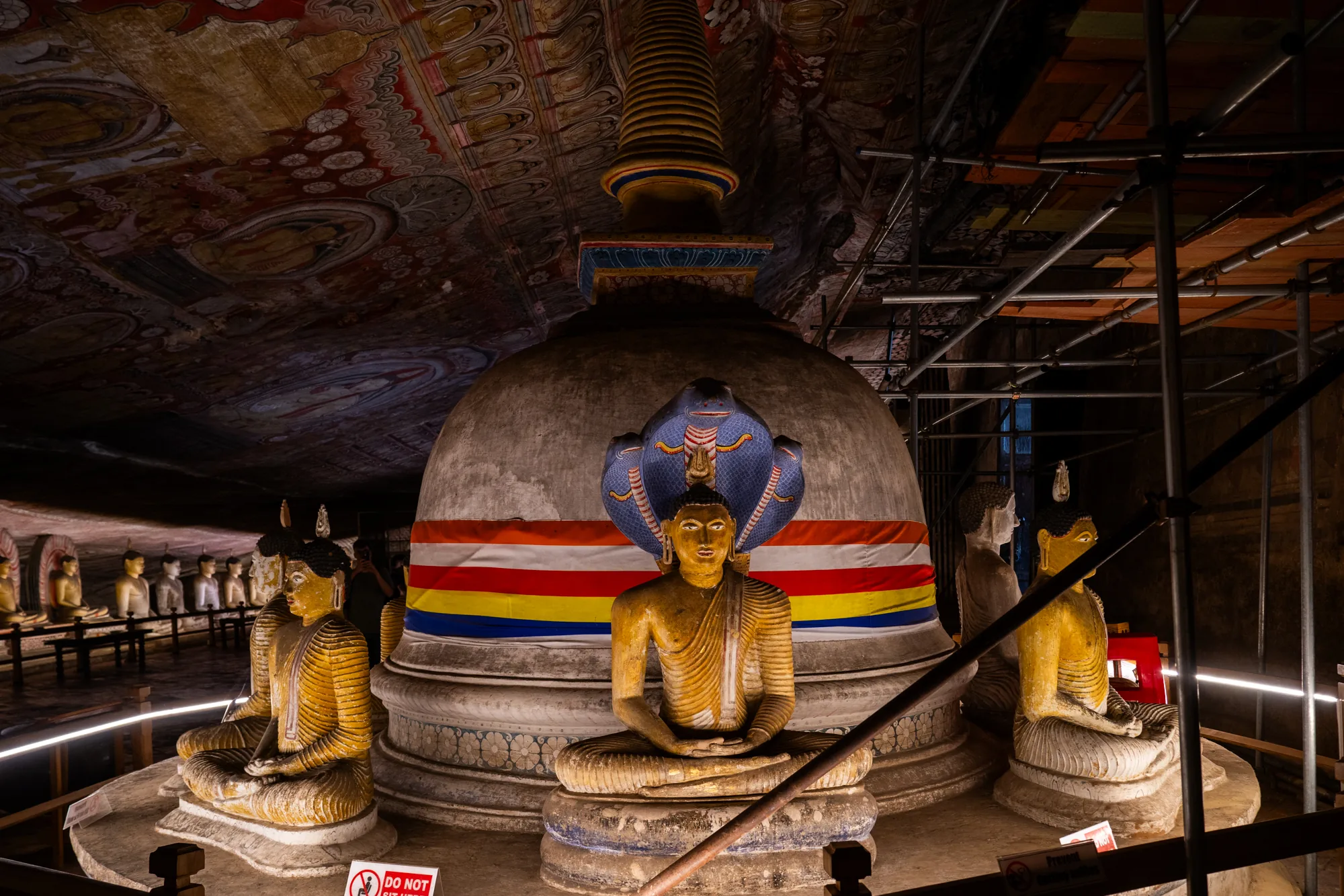 スリランカで撮影された、洞窟寺院内部の写真です。中央には大きな白い仏塔があり、その前には黄金色の仏像が座禅を組んでいます。仏塔にはスリランカ国旗の色が使われた帯が巻かれ、仏像の背後にはコブラの像が配置されています。背景には、洞窟の天井に描かれた壁画が見えます。壁画は人物や幾何学模様で彩られており、長い年月を経た古めかしい雰囲気を醸し出しています。写真全体はやや暗い照明で、神聖で落ち着いた雰囲気を強調しています。修復工事が行われているようで、画面右側には足場が組まれています。左側には一列に並んだ仏像が並び、その光景は神秘的です。
