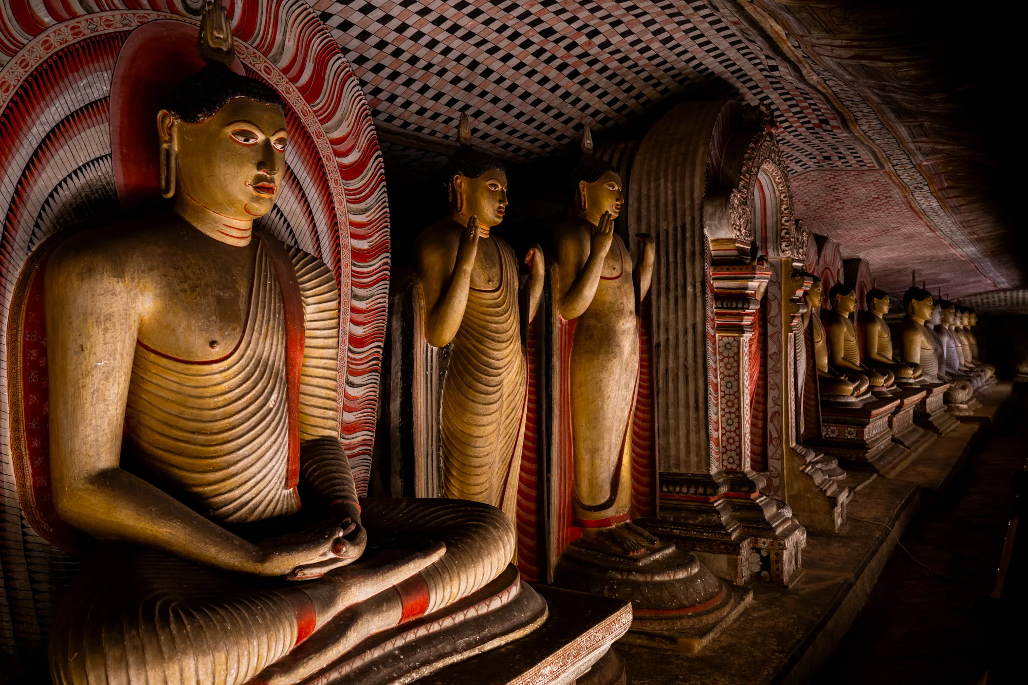 写真はスリランカで撮影されたもので、多数の仏像が並ぶ様子を捉えています。一番手前の仏像は座禅を組んだ姿で、穏やかな表情をしています。その背後には、立像の仏像が複数並んでおり、天井は幾何学模様で装飾されています。全体的に薄暗い照明で、仏像の荘厳さを際立たせています。赤と白の装飾が施されており、仏像の輪郭を強調し、深みのある空間を演出しています。
