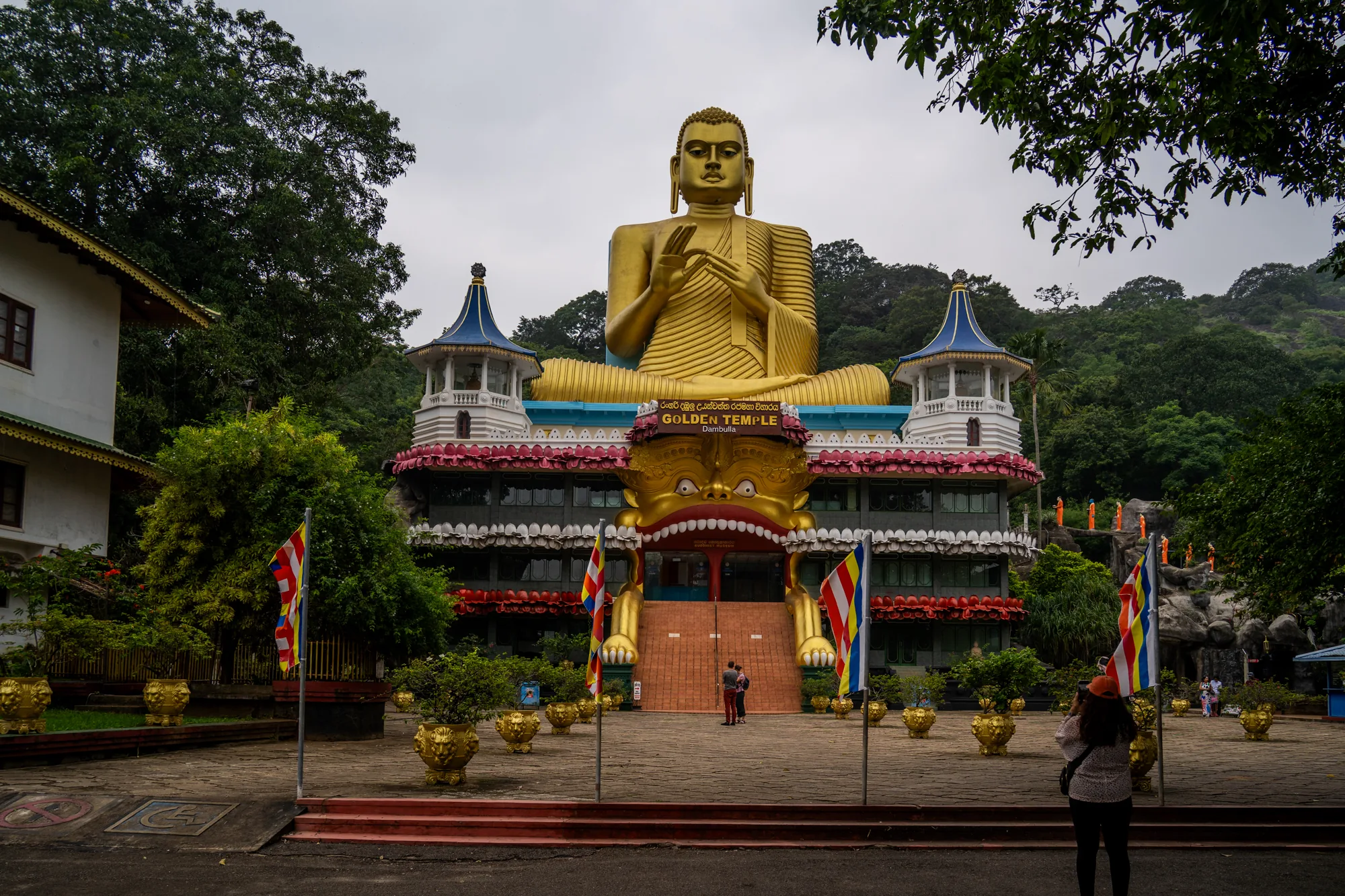 スリランカで撮影された写真です。中央には、大きな金色の仏像が鎮座する建物が写っています。建物の正面入り口は大きな口を開けた顔のデザインで、その両脇にはスリランカの国旗が立っています。入り口前にはカップルが立っており、建物の手前には黄金色の鉢植えが並べられています。画面右側には、カメラを構える女性の後ろ姿が見えます。周囲は緑豊かな木々に囲まれており、曇り空が厳かな雰囲気を醸し出しています。写真全体から、スリランカの宗教的な場所の壮大さが伝わってきます。また、画像の一部から、この寺院がダンブッラの黄金寺院であることが分かります。
