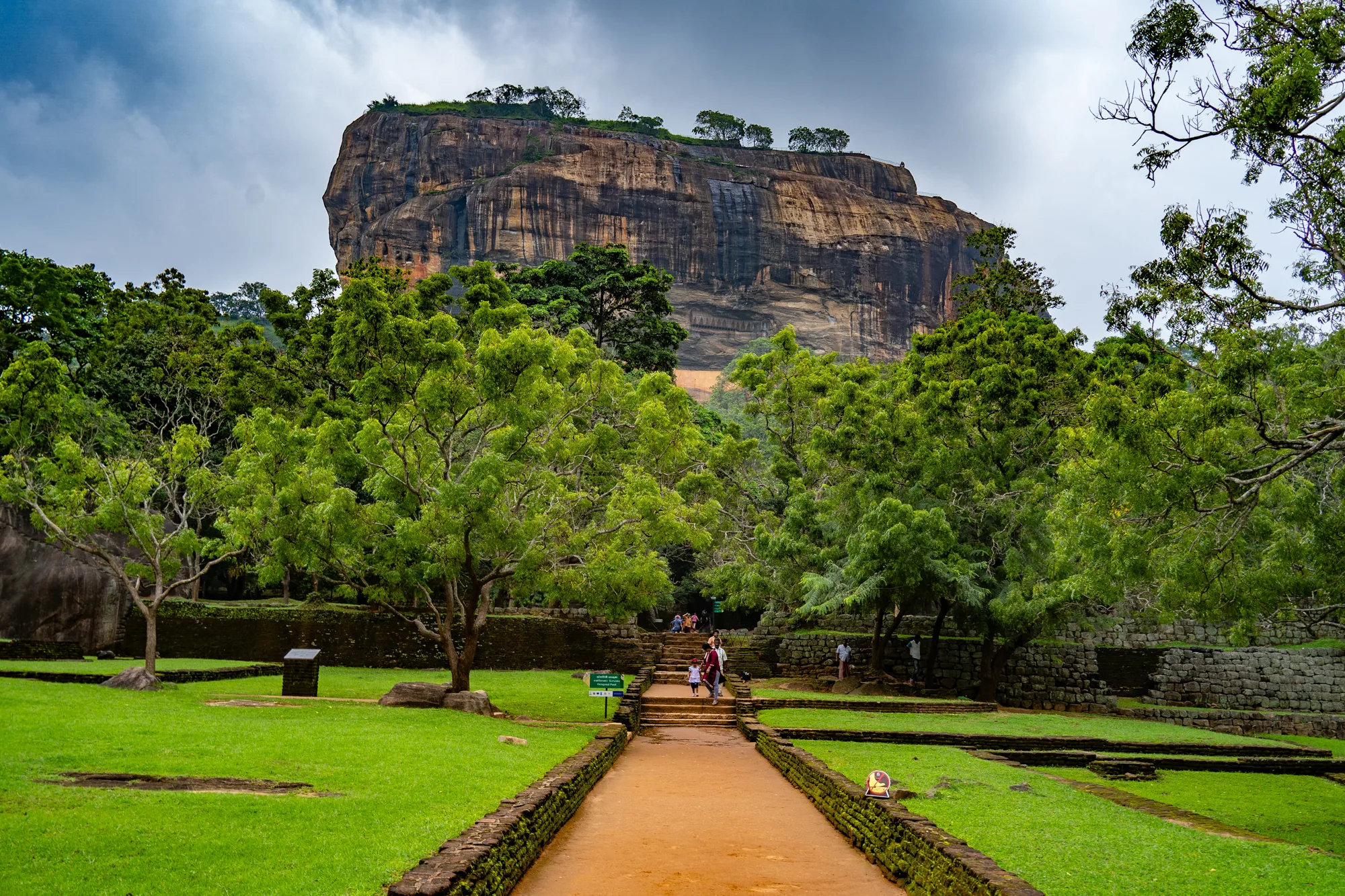 スリランカで撮影された、シーギリヤロックを捉えた写真です。画面奥には巨大な岩山がそびえ立ち、頂上には木々が生い茂っています。岩肌には雨水が流れた跡が見え、その歴史を感じさせます。岩山の手前には緑豊かな庭園が広がっており、中央には赤土の道が奥へと続いています。道沿いには石垣が築かれ、その一部には苔が生えています。庭園内には芝生が広がり、ところどころに遺跡のような石組みが見られます。道を進む人々も写っており、観光地であることを示唆しています。空は厚い雲に覆われていますが、全体的に明るい色調で、自然の雄大さを感じさせる写真です。