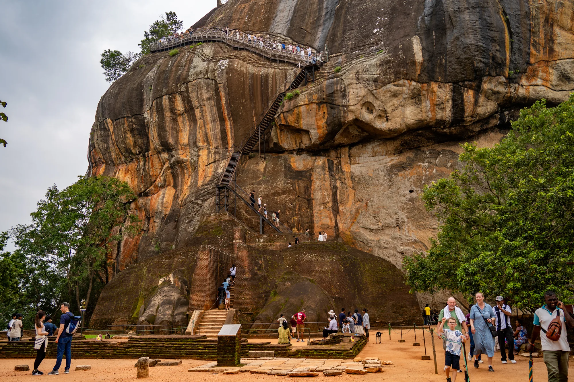 写真には、スリランカのシーギリヤロック（獅子岩）が大きく写っています。巨大な岩山は、赤茶色と灰色が混ざり合った独特の色合いをしており、その表面は風雨にさらされた歴史を感じさせる模様を描いています。岩山の頂上付近には、多くの観光客がおり、頂上へと続く鉄製の階段が岩肌に沿って設置されているのが見えます。階段を上る観光客も複数写っており、岩山への観光客の多さを物語っています。  岩山の麓には、古代の遺跡と思われるレンガ造りの構造物が残っており、その上に生えた苔が歴史の重みを感じさせます。麓の地面は赤土で覆われており、多くの観光客が散策しています。様々な人種の観光客が写っており、シーギリヤロックが世界的な観光地であることがわかります。画面右側には緑豊かな木々が生い茂り、自然と遺跡が調和した美しい風景を作り出しています。空は薄曇りで、全体的に落ち着いた雰囲気の写真です。