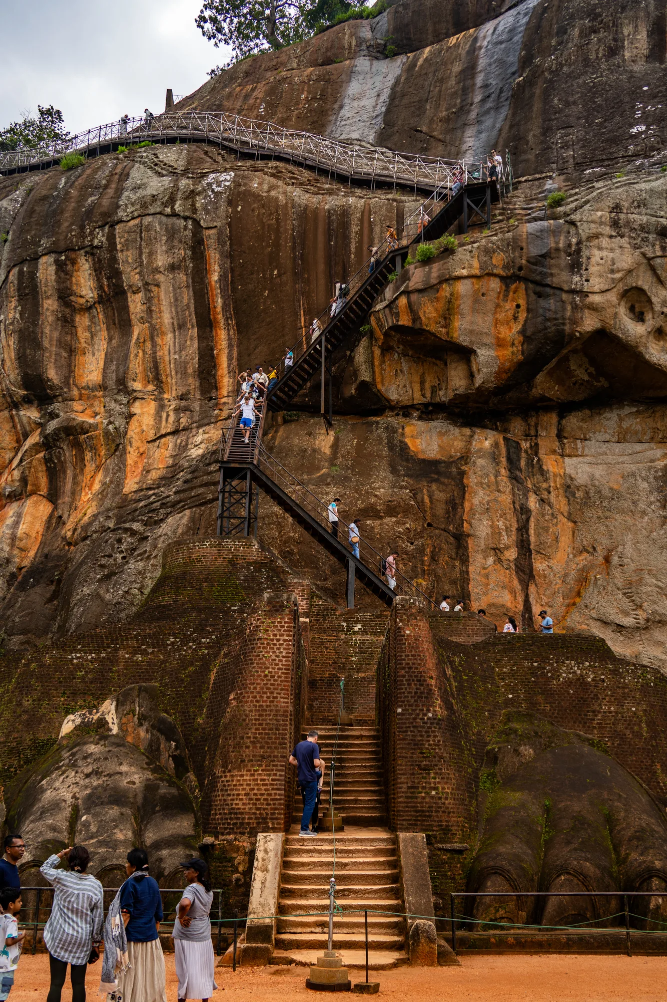 スリランカで撮影された写真には、観光客がシーギリヤロックの頂上を目指して階段を上る様子が写っています。画面全体を覆うように巨大な岩山がそびえ立ち、その表面はオレンジ色と灰色が入り混じった独特の色合いを見せています。岩山には、赤茶色のレンガで作られた構造物や、頂上へと続く金属製の階段が設置されています。階段には多くの観光客がおり、それぞれのペースで頂上を目指しています。岩山の頂上には木々が生い茂っており、空は曇りがちです。階段のふもとには観光客が集まっており、これから階段を上る人もいれば、休憩している人もいます。地面は赤土で覆われており、周囲の景色と調和しています。全体的に、写真からはシーギリヤロックの壮大さと、それを訪れる観光客の熱気が伝わってきます。