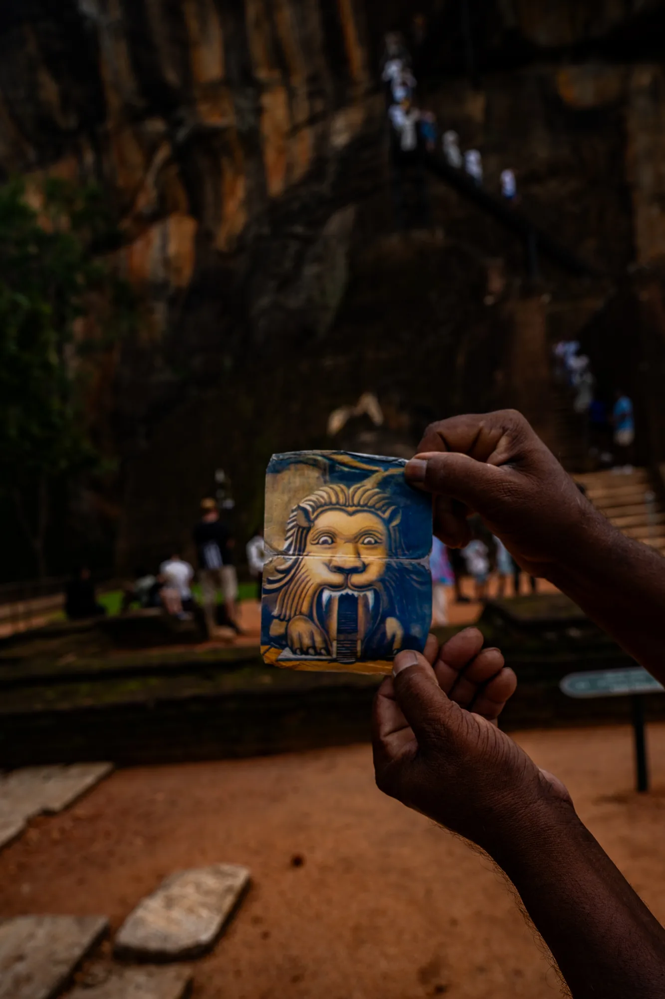 スリランカのシーギリヤロック付近で撮影された写真です。前景には、男性の手がライオンの口を模した遺跡の絵が描かれたタイルを持ち上げています。絵のライオンは、入り口のような口を開けており、シーギリヤロックにあるライオンの門を連想させます。背景には、そびえ立つ岩壁と、岩肌に取り付けられた階段を登る人々の姿が見えます。足元は赤土で覆われており、観光客らしき人々が散見されます。全体的に、歴史的な遺跡と観光地の雰囲気が感じられます。