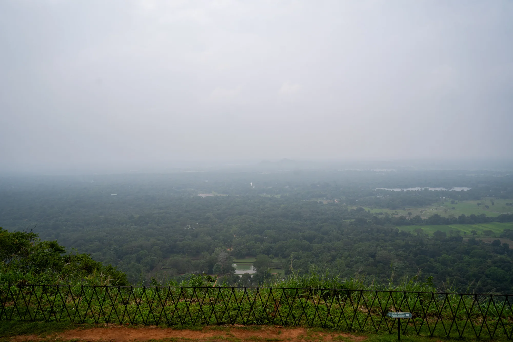 写真はスリランカで撮影されたもので、高い場所から見下ろす風景が広がっています。全体的に霧がかっており、遠くの景色はぼやけて見えます。手前には柵があり、その向こうには緑豊かな森林が広がっています。森林の中には、小さな湖や池のような水面が見えます。遠景は霧に覆われていますが、かすかに山のような地形が見て取れます。全体的に落ち着いた雰囲気で、霧が幻想的な雰囲気を醸し出しています。手前の柵と、足元の土の質感から、安全な場所から景色を眺めている様子が伝わってきます。