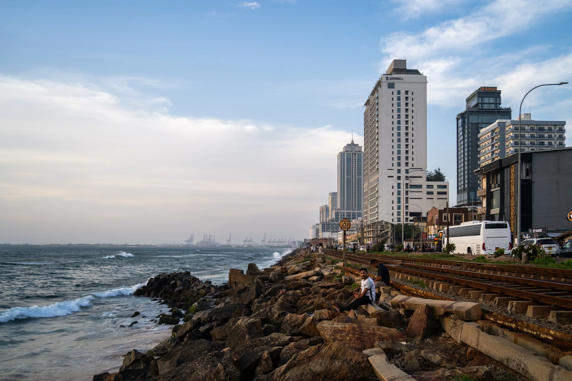 写真はスリランカで撮影されたもので、海岸沿いの都市風景が写っています。手前には岩場があり、波が打ち寄せています。岩場には二人の男性が座って休憩している様子が見えます。岩場のすぐ隣には線路が敷かれており、その奥には道路と建物が並んでいます。高層ビルもいくつか見え、都市部であることを示唆しています。遠くには港湾施設のようなものが見え、海運が盛んな地域であることが伺えます。空は晴れており、薄い雲が広がっています。全体的に落ち着いた雰囲気の写真です。