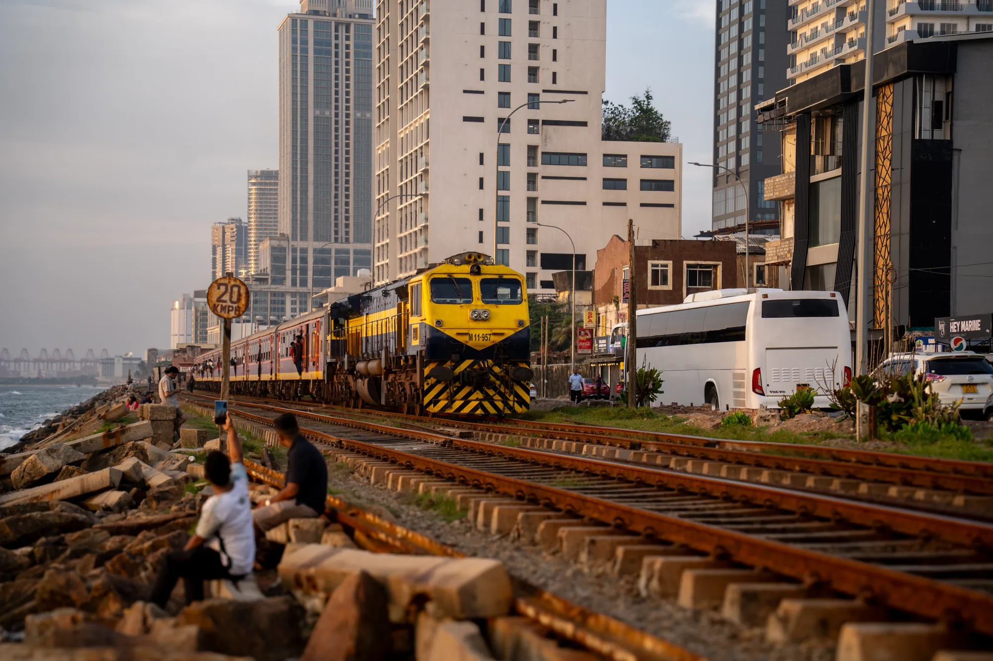 スリランカで撮影されたと思われる写真です。手前には鉄道の線路が左右に伸び、その横には大きな岩が積み重ねられた海岸線が見えます。海岸線沿いには人が数人座っており、一人はスマートフォンを高く掲げて写真を撮っているようです。奥には黄色と黒のディーゼル機関車が牽引する列車が写っており、乗客が窓から顔を出しています。列車の背景には、高層ビル群が立ち並ぶ都市の景観が広がっています。画面右側には白いバスや建物の一部が見え、都市の生活感を感じさせます。全体的に夕暮れ時の柔らかな光が差し込み、穏やかな雰囲気を醸し出しています。線路脇の制限速度標識には「20 KMPH」と記載されています。
