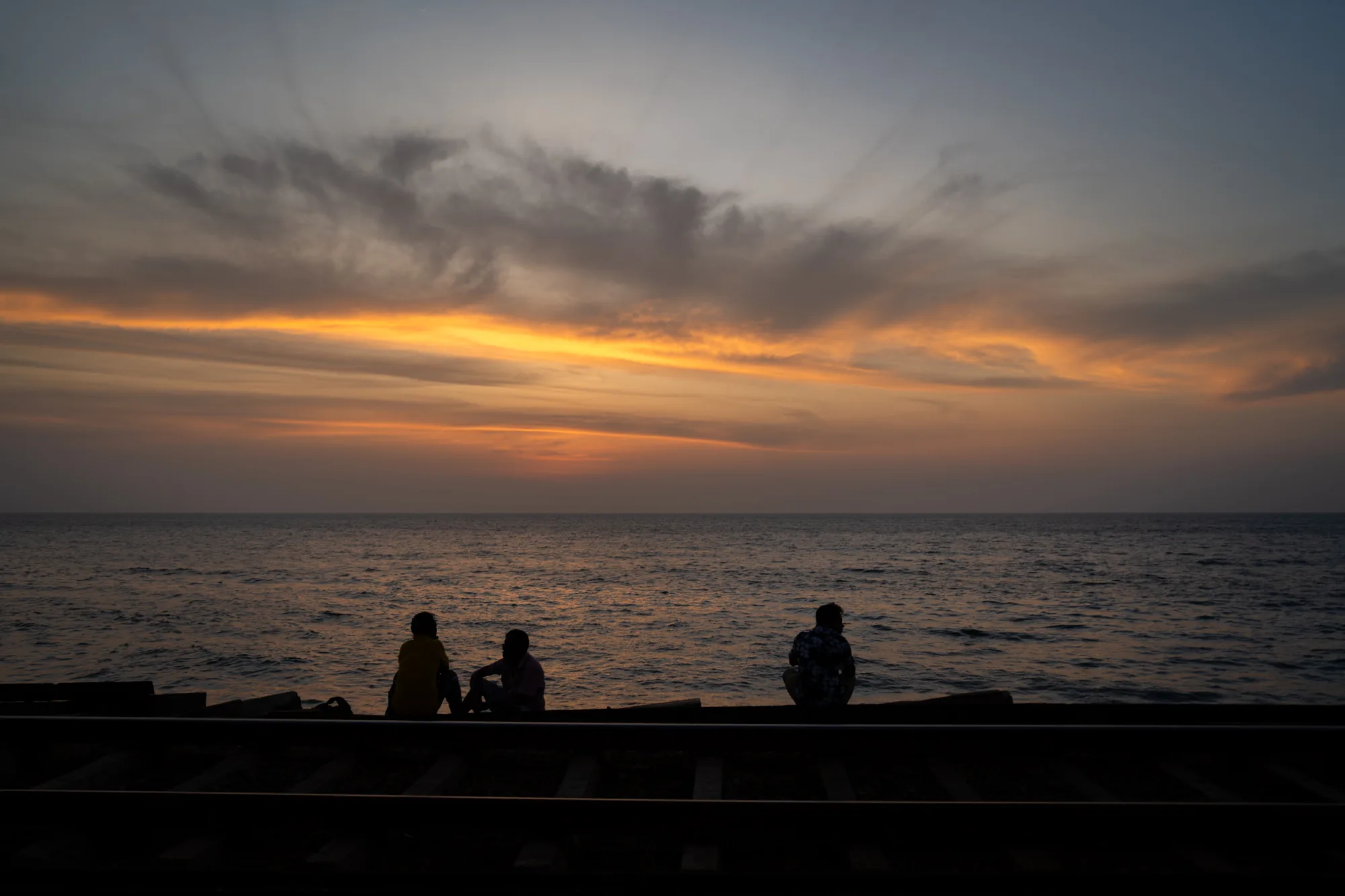 スリランカで撮影されたと思われるこの写真は、美しい夕焼けを背景に、海岸沿いの線路に座る人々のシルエットが印象的です。空は上部が薄い青色から、水平線に向かって鮮やかなオレンジ色にグラデーションを描いています。雲はたなびき、夕日の光を受けて金色に輝いています。海は穏やかで、夕焼けの色を反射してきらめいています。前景には線路があり、その上に3人の人物が座っています。彼らはシルエットになっており、詳細は分かりませんが、リラックスした様子で海を眺めているように見えます。全体の雰囲気は静かで穏やかで、瞑想的な気分に誘われます。