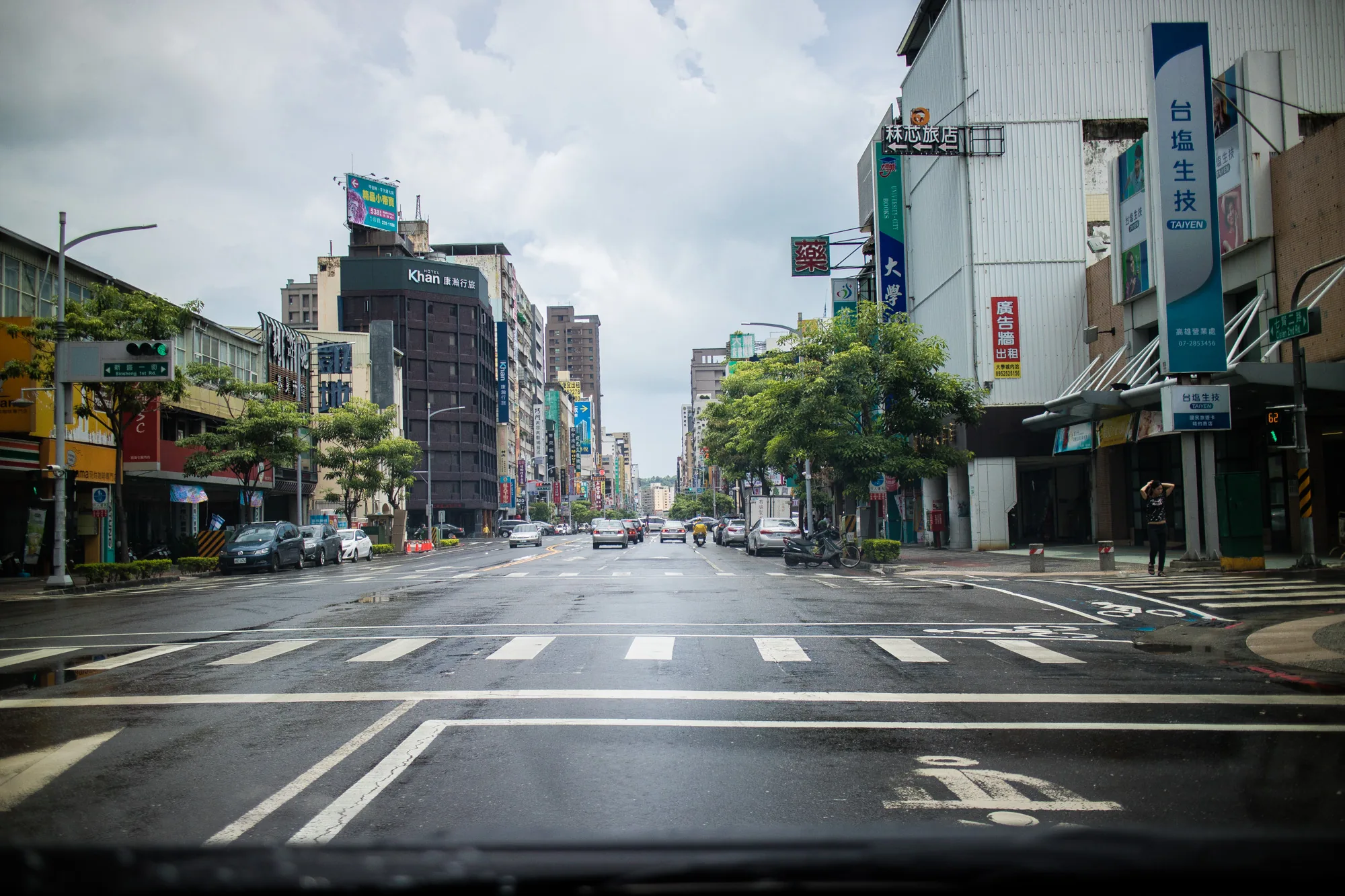 この写真は台湾の台南で撮影されたもので、雨上がりの道路が濡れて光を反射しているのが印象的です。視点は車の運転席からのようで、フロントガラスの下部がわずかに写り込んでいます。画面中央には、まっすぐに伸びる道路があり、左右には建物が立ち並んでいます。  建物の多くは広告看板で覆われており、様々な言語の文字やロゴが見て取れます。特に「Khan 康瀚行旅」というホテルの看板や、「台塩生技 TAIYEN」という企業の広告が目立ちます。また、林芯旅店、UNIVERSITY-CITY BOOKS大学などの看板も見られます。  道路には車やバイクが走行しており、歩道には人が歩いています。信号機も設置されており、交通整理が行われている様子がわかります。空は曇っており、全体的に落ち着いた雰囲気の写真です。道路の白線や横断歩道もはっきりと描かれており、都市部の日常風景を切り取ったような印象を受けます。