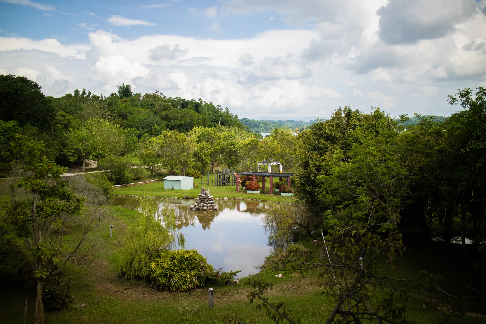 台湾の台南で撮影されたと思われる写真です。緑豊かな庭園風景が広がっています。写真中央には池があり、水面には空の雲が鏡のように映り込んでいます。池の中央には石を積み重ねたオブジェがあり、その周りには草木が生い茂っています。池の奥には、休憩所のような構造物が見えます。また、青い物置小屋や、子供用の遊具らしきものも見受けられます。庭園全体を囲むように木々が生い茂り、遠景にはうっすらと山並みが見えます。空には白い雲が浮かび、穏やかな雰囲気を醸し出しています。全体的に手入れが行き届いた庭園であり、静かで落ち着いた空間であることがうかがえます。