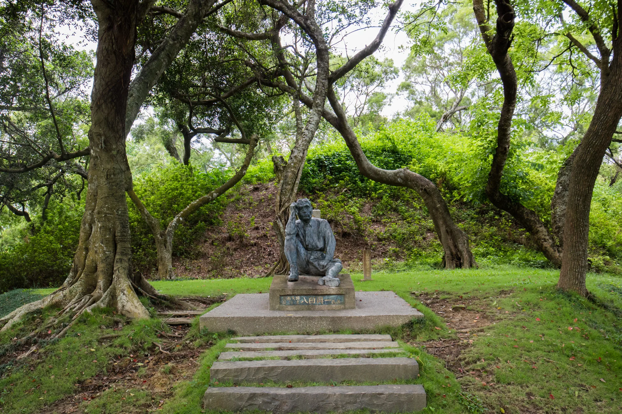 台湾の台南で撮影された写真です。緑豊かな公園の中に、石段を上がった場所に、男性の彫像が置かれています。彫像は何か考え込んでいるようなポーズをしており、落ち着いた雰囲気です。彫像の背後には緑が生い茂り、木々が木陰を作っています。石段の周りも緑で覆われており、自然と調和した静寂な空間が広がっています。全体的に落ち着いたトーンで、穏やかな時間が流れているような印象を受けます。