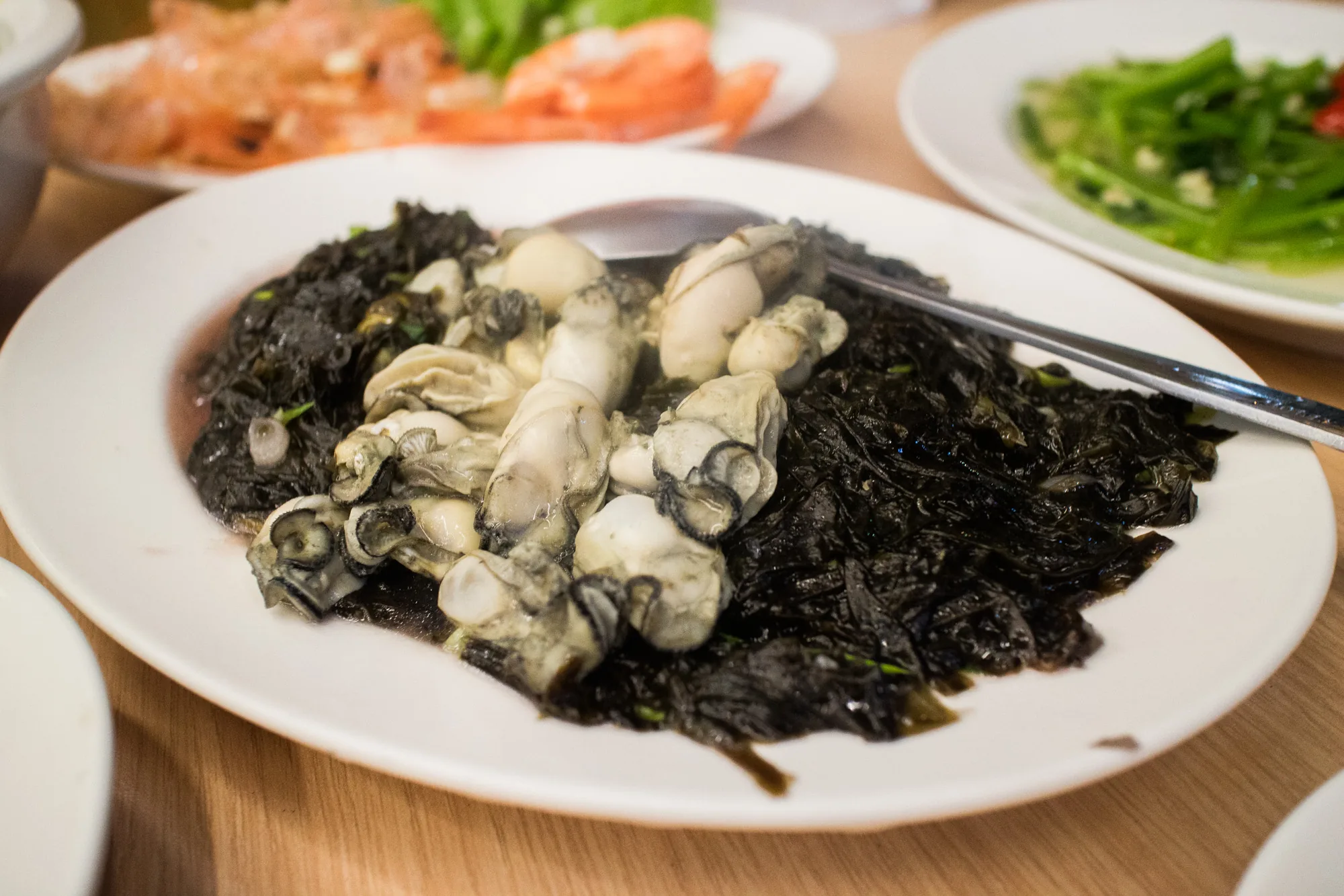 台湾の台南で撮影された写真です。木製のテーブルの上に白い皿が置かれ、皿には牡蠣と海苔の炒め物のような料理が盛り付けられています。牡蠣はふっくらとしていて新鮮そうで、海苔は照りがあり、醤油ベースのタレで炒められているようです。皿の横には取り分け用のトングが添えられています。奥にはエビチリのような料理や、青菜の炒め物のような料理が写っており、賑やかな食卓の様子が伺えます。全体的に美味しそうな雰囲気で、台湾料理への食欲をそそる写真です。