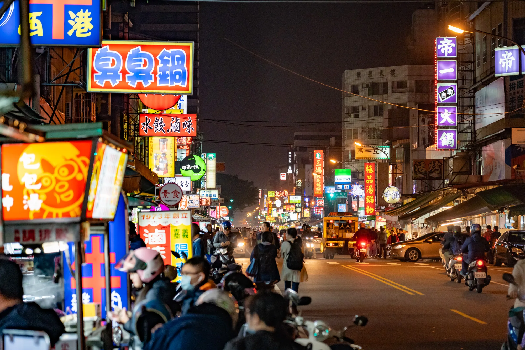 写真は台湾の台北で撮影された夜の街並みを捉えています。通りには多数のネオンサインが輝き、日本語、中国語、英語などの文字が見て取れます。特に飲食店の看板が多く、台湾料理店の名前が目立ちます。「臭臭鍋」という看板が複数あり、台湾名物料理のようです。  通りは人通りが多く、多くの人が歩いています。特にスクーターに乗った人々が多く、台北の交通手段としてスクーターが一般的であることが伺えます。車も走っており、タクシーも確認できます。  写真全体は夜の賑やかな雰囲気を捉えており、台北の活気ある様子が伝わってきます。