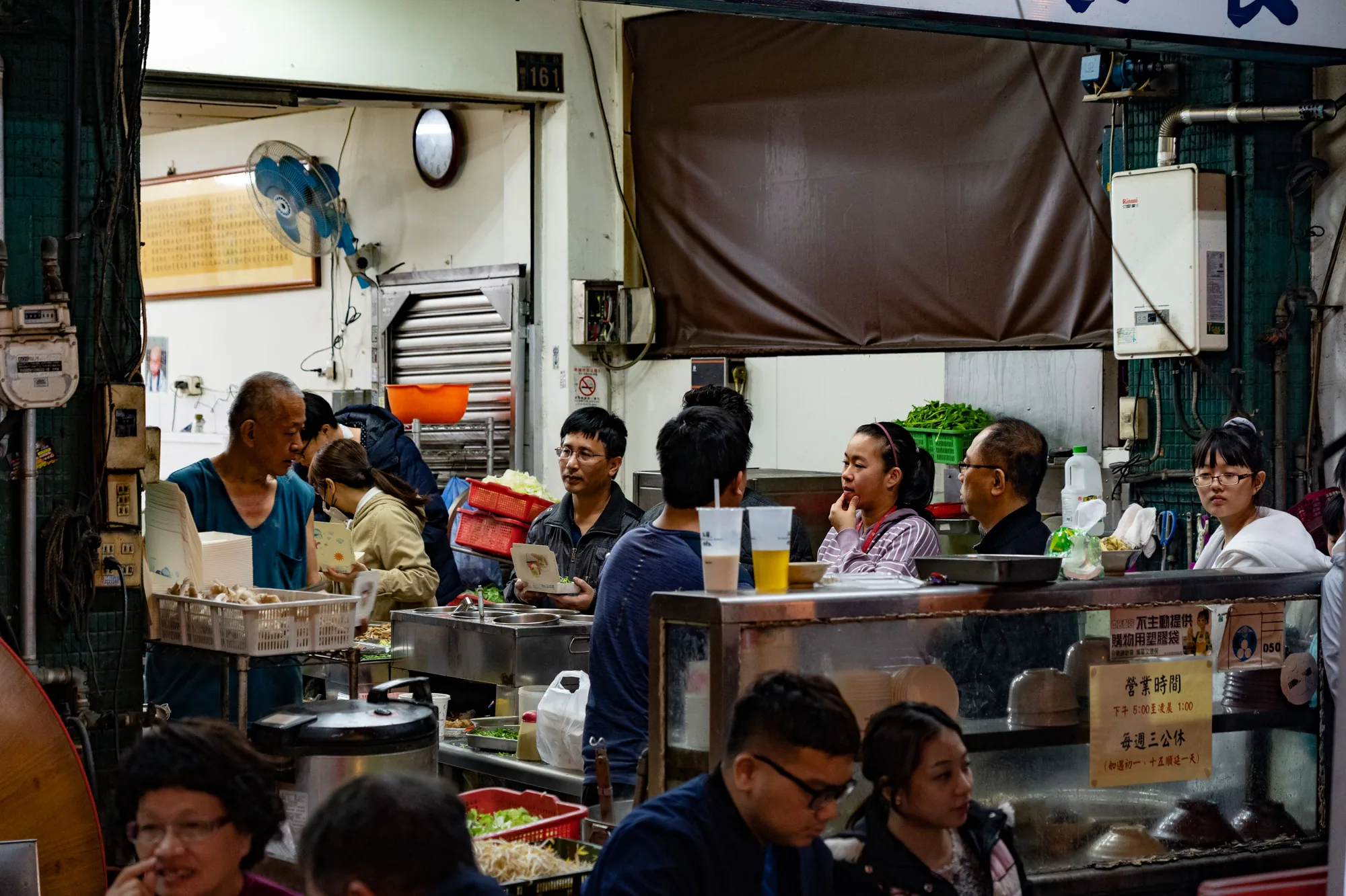 写真は、台湾の台北で撮影された屋台料理店の様子を捉えたものです。店は活気にあふれており、多くの人々が食事を楽しんでいます。手前には料理を待つ客が座っており、奥には調理場が見えます。調理場では、店員が忙しそうに料理を作っています。  調理場には、様々な食材や調理器具が並んでいます。新鮮な野菜や肉、麺類などが見られます。店員は手際よくこれらの食材を調理し、客に提供しています。店内の壁には、メニューや営業時間が書かれた看板が掲げられています。  店内の雰囲気は賑やかで、活気に満ち溢れています。人々は料理を味わいながら談笑し、楽しい時間を過ごしています。写真は、台北の屋台料理店の日常をリアルに切り取った一枚と言えるでしょう。