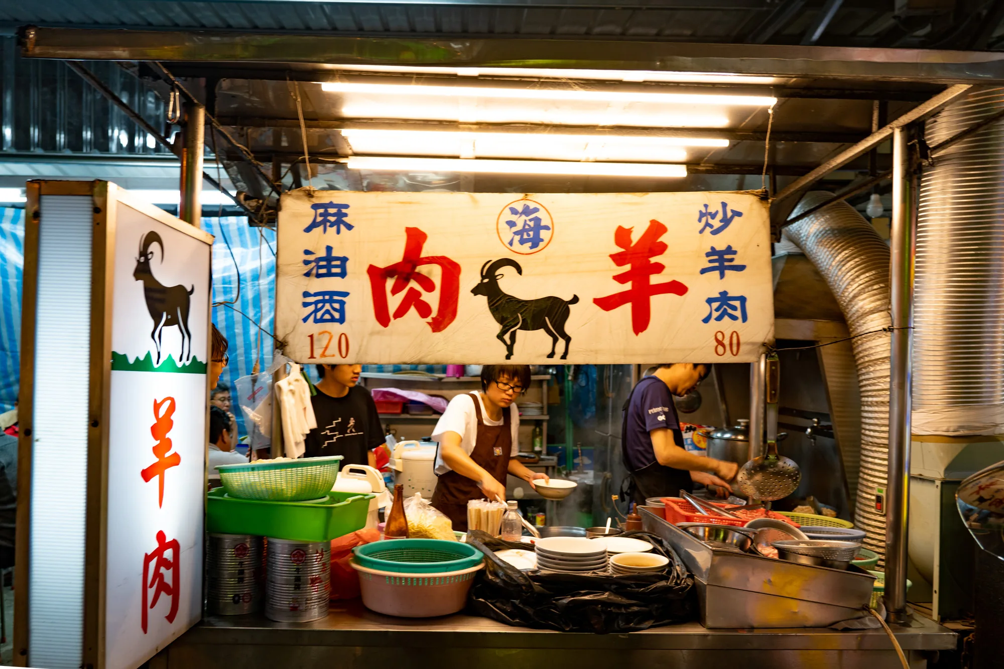 写真は台湾の台北にある羊肉料理店の様子を捉えています。店先には「麻油酒肉 120」、「海羊」、「炒羊肉 80」と書かれた看板が掲げられており、価格が表示されています。看板には羊のシルエットが描かれています。調理台では、エプロンをかけたスタッフが料理を調理しており、もう一人のスタッフが調理の準備をしています。調理台には、調理器具や食材が並べられ、活気のある雰囲気が伝わってきます。店構えは簡素であり、台湾のローカルな雰囲気が感じられます。蛍光灯の光が料理や看板を照らし出し、食欲をそそるような印象を与えています。