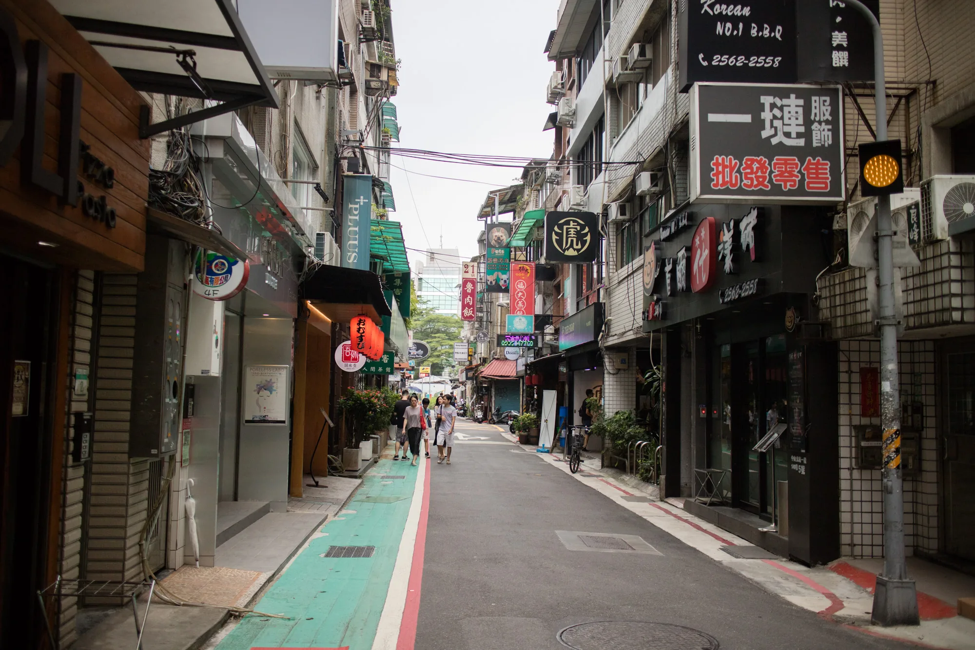 写真は台湾の台北で撮影された、賑やかな路地裏の様子を捉えています。路地はやや狭く、両側には様々な店舗が軒を連ねています。店先にはカラフルな看板や提灯が掲げられ、アジアらしい活気ある雰囲気を醸し出しています。通りには数人の歩行者がおり、日常の風景の一部を構成しています。路面はアスファルトで、歩道部分は緑色に塗られています。写真全体はやや曇りがかった空の下で撮影されており、落ち着いた色調です。電線が空中に張り巡らされ、都市の生活感を強調しています。飲食店の看板が多く、現地の食文化を垣間見ることができます。