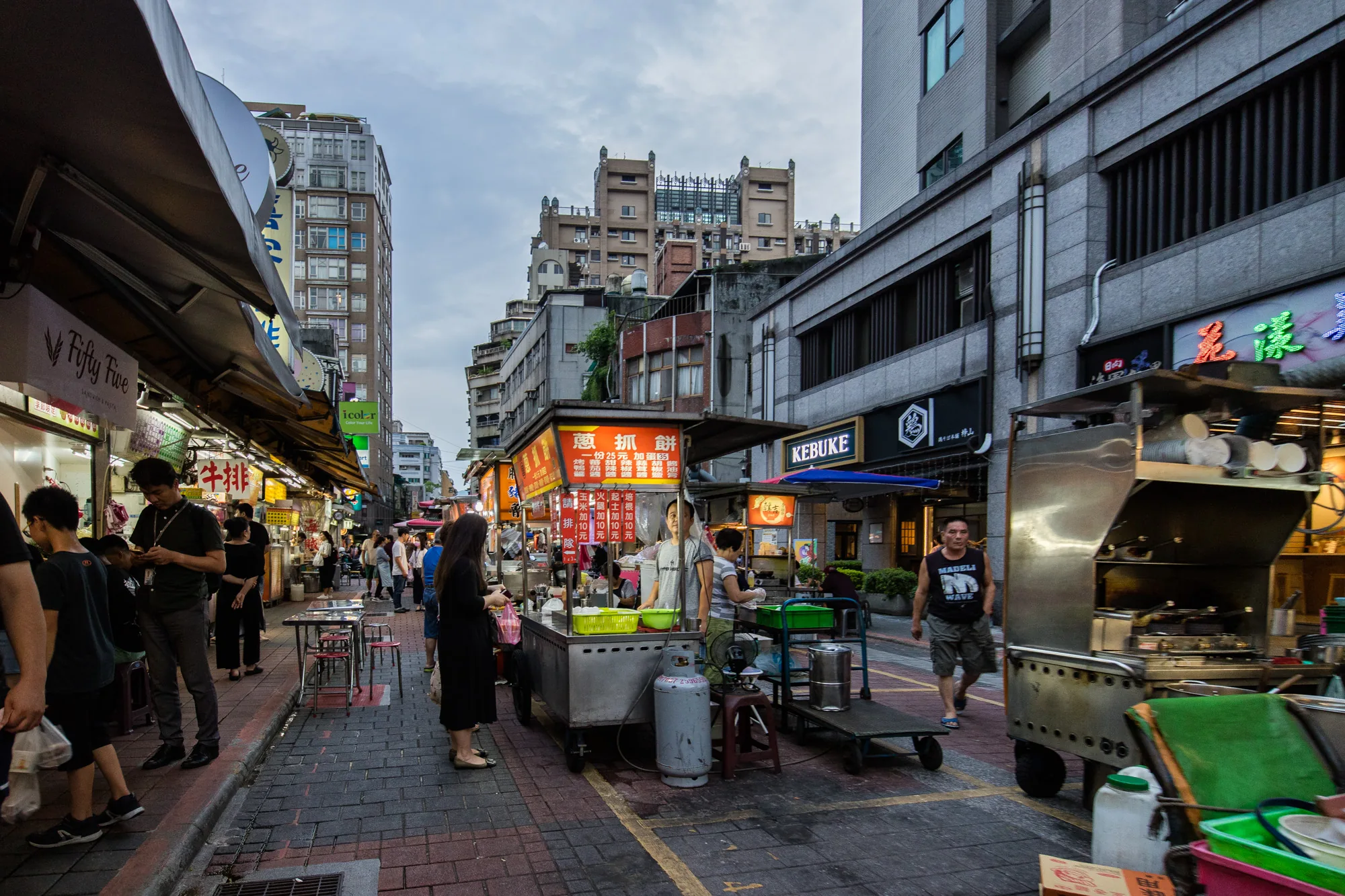 この写真は、台湾の台北にある賑やかな屋台街の様子を捉えています。夕暮れ時の空の下、レンガ敷きの通りには、様々な屋台が軒を連ね、多くの人々で賑わっています。  左側には、軒先に店名が書かれた看板が並び、多くの人が食べ物を買い求めています。看板には「Fifty Five SANDWICH & PASTA」や「牛排」といった文字が見えます。路上には、テーブルと椅子が置かれ、人々が食事を楽しんでいる様子もうかがえます。  中央には、屋台の屋根に「蔥抓餅」と書かれた、ひときわ目立つ屋台があります。ここでは、葱抓餅（ネギパンケーキ）を販売しているようです。屋台の前には、注文を待つ人々が列を作っています。その隣には、「KEBUKE」と書かれた看板が見えます。  右側には、ステンレス製の屋台が見え、その横を男性が歩いています。屋台の奥には、高い建物が立ち並び、都会の風景が広がっています。空は曇り空で、夕暮れ時の柔らかな光が、街全体を包み込んでいます。  全体的に、活気と賑わいのある、台北の屋台街の日常風景を切り取った写真と言えるでしょう。様々な食べ物の香りや人々の話し声が聞こえてくるような、臨場感あふれる一枚です。