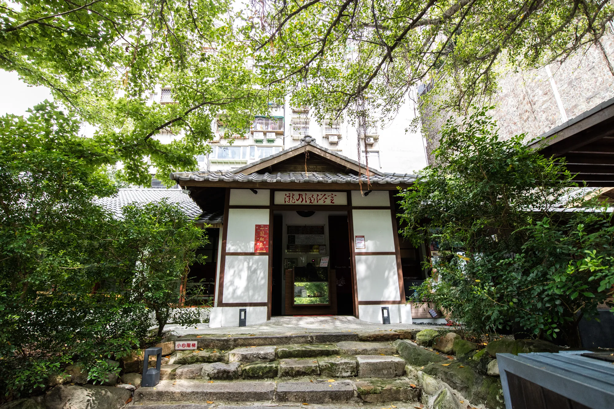 写真は、台湾の台北にあると思われる和風建築物を正面から捉えたものです。石段を上った先に建物があり、周囲は緑の木々や茂みで覆われています。建物は白壁に茶色の枠組みが特徴で、屋根は瓦葺きです。入り口の上には「瀧乃湯浴室」と書かれた看板が見えます。建物の奥には、現代的な建物も写っており、新旧の建物が混在する風景が印象的です。石段には苔が生えており、歴史を感じさせます。手前には「小心階梯」と書かれた注意書きの看板があります。全体的に、静かで落ち着いた雰囲気の写真です。