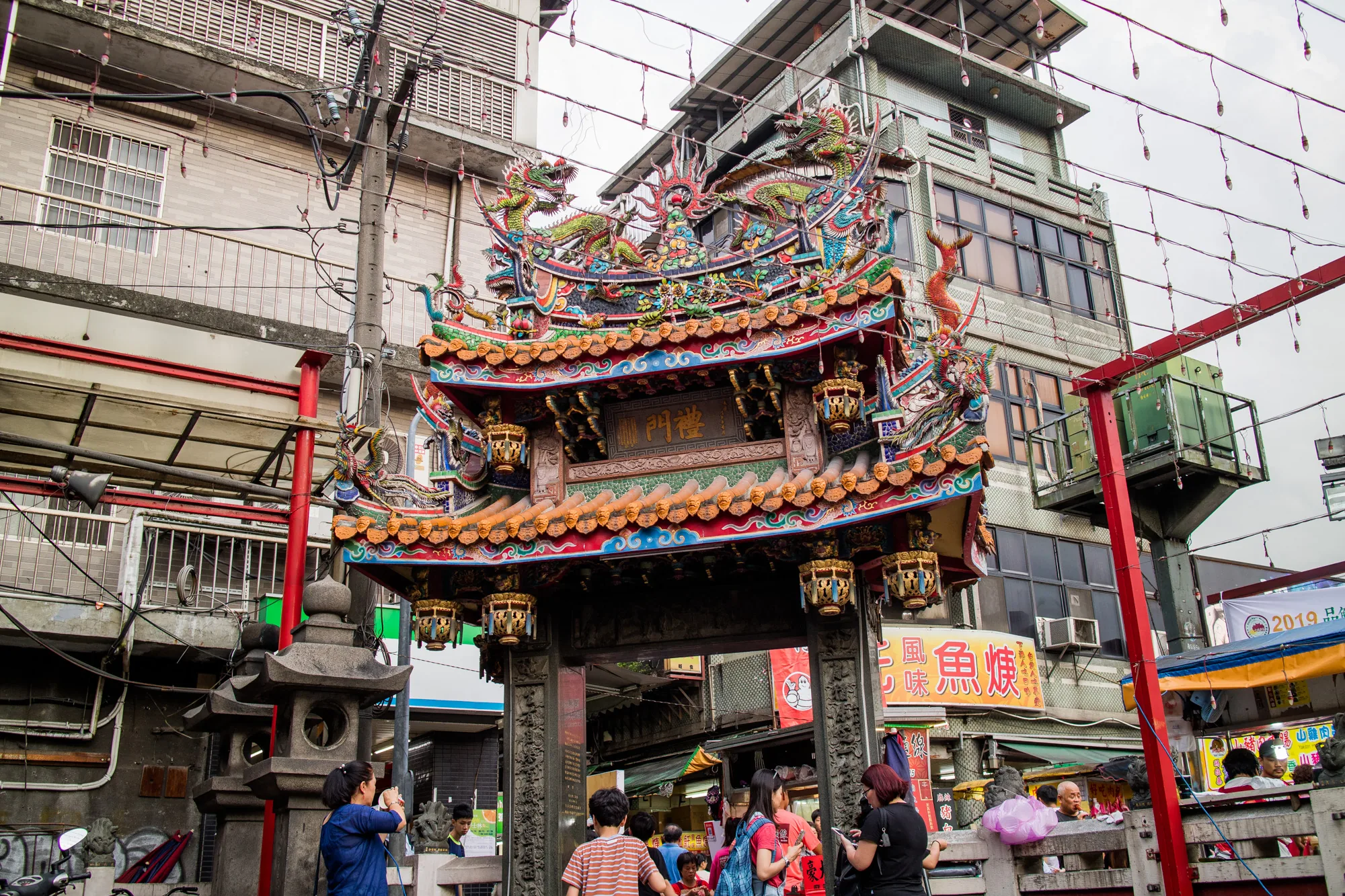 写真は台湾の台北で撮影されたもので、伝統的な装飾が施された寺院の門が中心に写っています。門の上部には、鮮やかな色彩の龍や鳳凰の彫刻が施され、細部まで丁寧に作り込まれています。門の奥には、賑やかな市場のような雰囲気の通りが広がっており、多くの人々が行き交っています。写真手前には、寺院の石灯籠があり、人々がスマートフォンで写真を撮っている様子も見られます。背景には、近代的な建物が立ち並び、伝統と現代が混ざり合った台北の風景を捉えています。全体的に、活気があり、文化的な魅力を感じさせる一枚です。