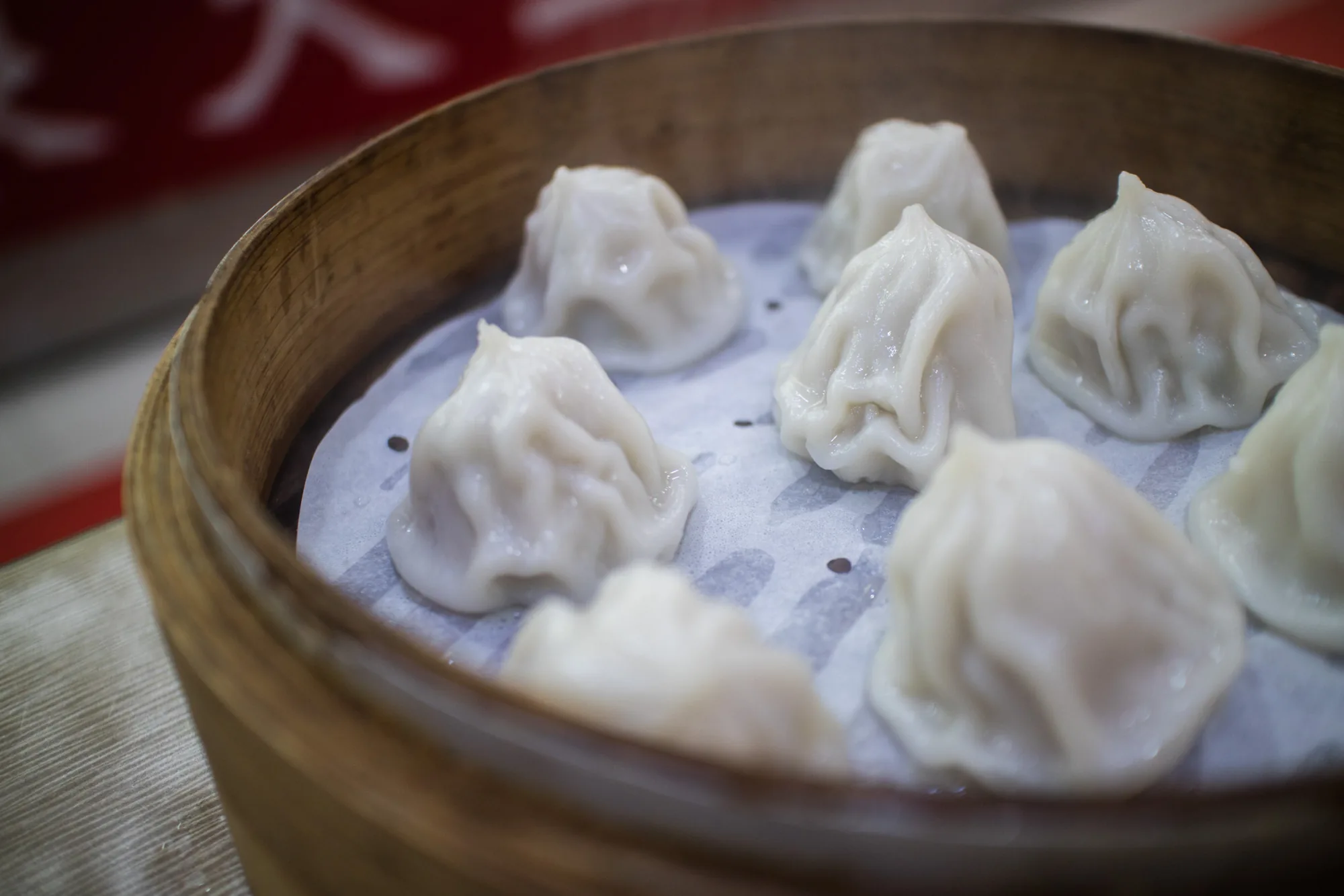 写真には、台湾の台北で撮影されたと思われる小籠包が写っています。木製の蒸篭に、クッキングシートが敷かれ、その上に白い、つややかな小籠包が7つ並んでいます。小籠包は、薄皮で包まれており、丁寧にひだを寄せて成形されています。蒸し上げられたばかりで、かすかに湯気が立っているようにも見えます。背景には、赤と白のぼかしが見え、お店の装飾の一部かもしれません。全体的に温かく、食欲をそそる雰囲気の写真です。