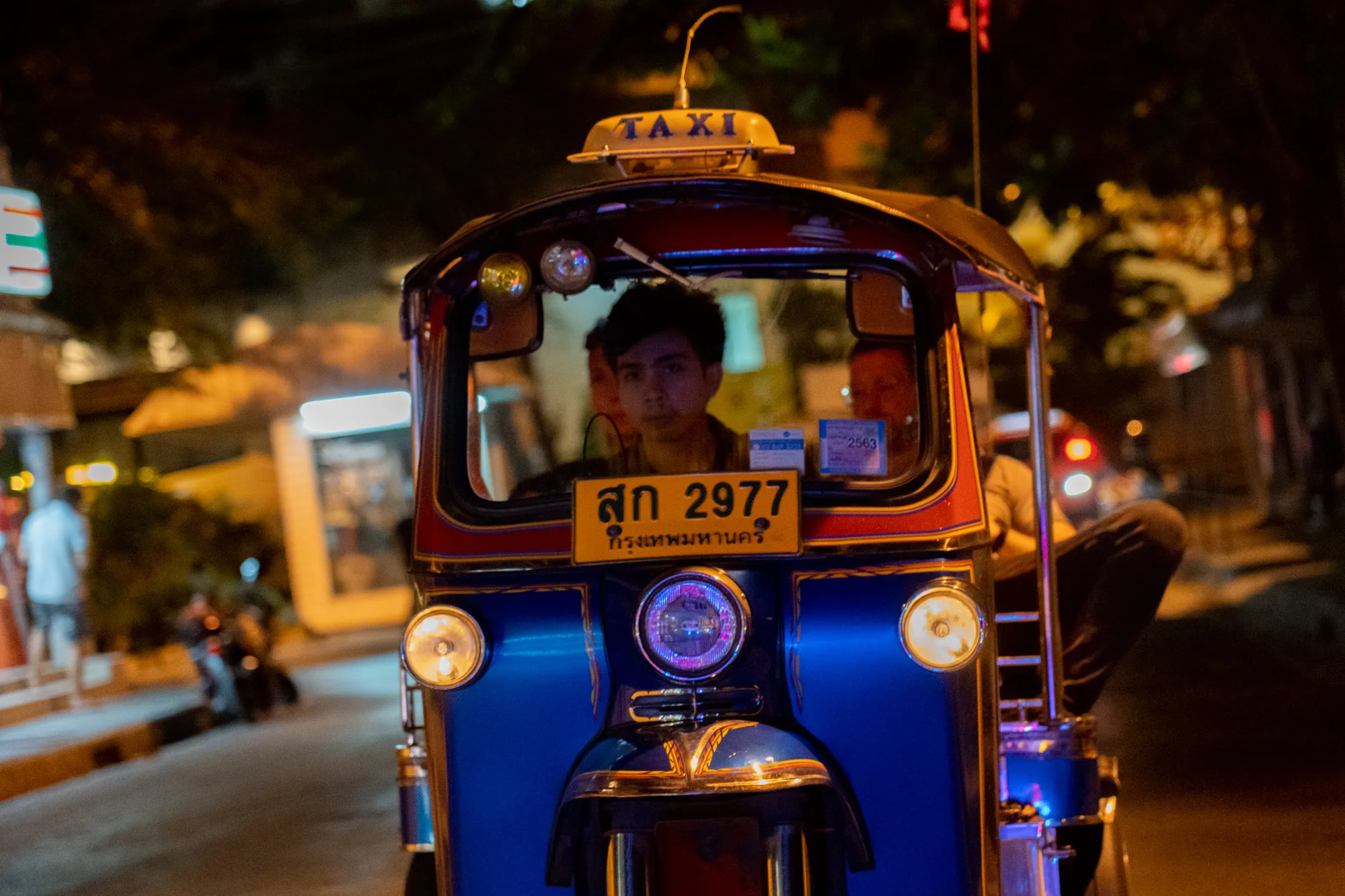 写真はタイのバンコクで撮影された、夜のトゥクトゥクの様子を捉えたものです。トゥクトゥクは鮮やかな青色で、赤色の縁取りが施されています。正面には「TAXI」の文字が入ったサインが付いています。運転手は若い男性で、真剣な表情で前を見ています。助手席には別の人も乗っているようです。トゥクトゥクのナンバープレートにはタイ語の数字が書かれています。周囲は夜の街並みで、背景にはぼやけた光や建物が見えます。街灯や他の車両のライトが点灯しており、賑やかな夜の雰囲気が伝わってきます。路面には光が反射しており、夜の街の湿気を感じさせます。写真全体を通して、エキゾチックなタイの雰囲気が漂っています。