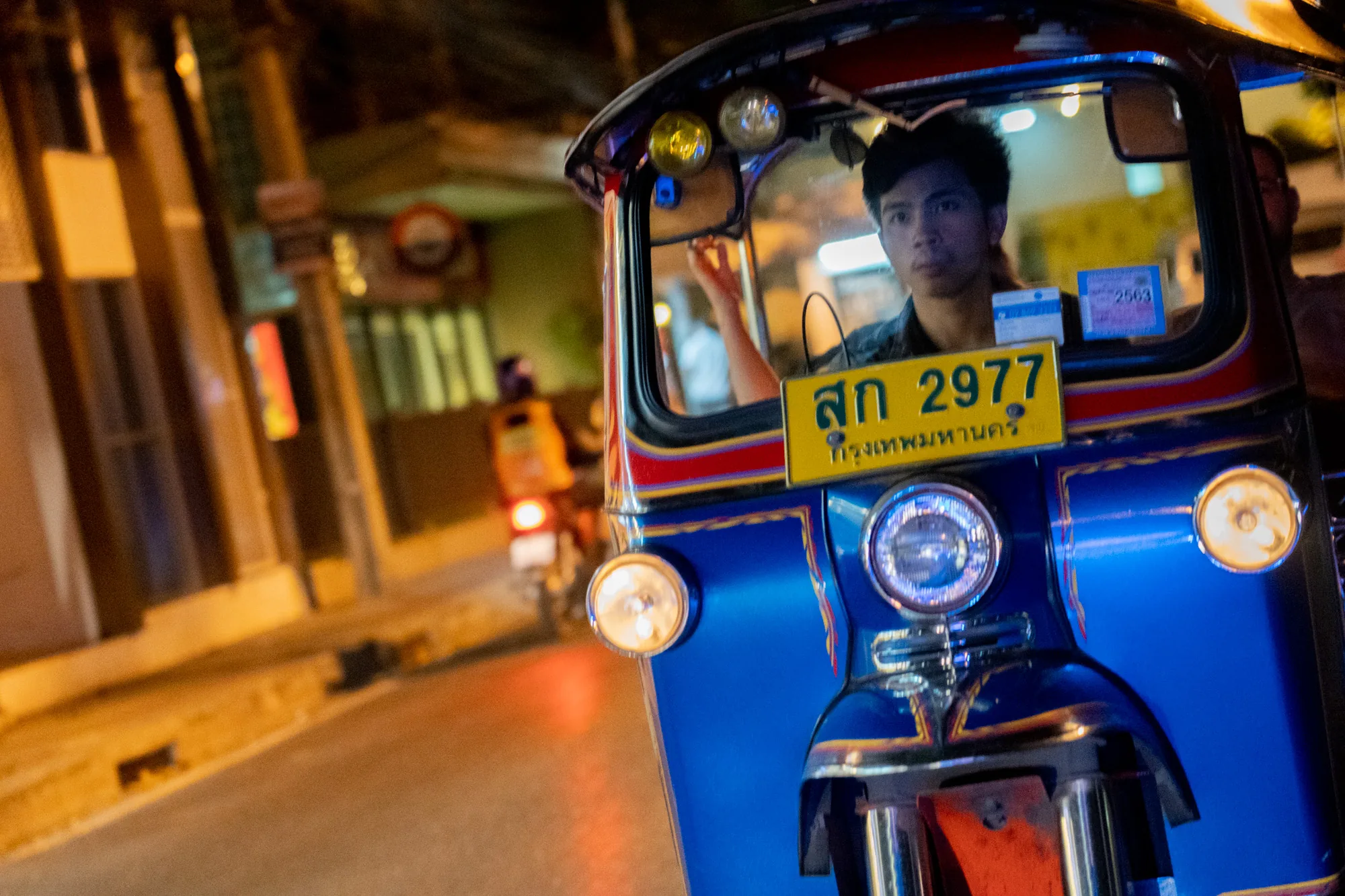 写真はタイのバンコクで撮影された夜の風景です。鮮やかな青色の車体のトゥクトゥクが正面を向いて写っており、運転手の若い男性がこちらを見ています。運転手は黒髪で、黒いシャツを着ています。トゥクトゥクのフロントには黄色いナンバープレートがあり、「สก 2977 กรุงเทพมหานคร」とタイ語で書かれています。ナンバープレートの上には小さな白いステッカーが貼られています。トゥクトゥクのヘッドライトは点灯しており、周囲を照らしています。背景はぼやけていますが、建物の明かりや街灯が写っており、夜の賑やかな様子が伺えます。トゥクトゥクの後ろにはバイクが止まっており、全体的に活気のある、アジアの夜の雰囲気が伝わってくる写真です。