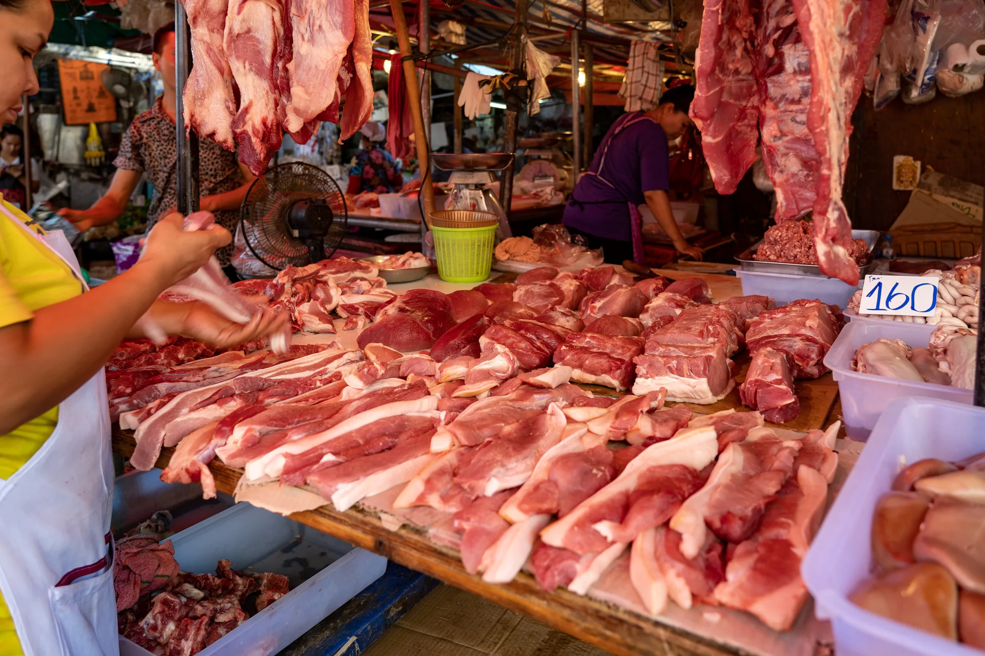 写真はタイのバンコクにある肉屋の店先の様子を捉えています。店の前には、様々な種類の肉が山積みにされています。手前には薄切りの豚肉が並び、奥には塊の肉やひき肉が置かれています。天井からは大きな肉塊が吊るされており、活気のある市場の雰囲気を醸し出しています。店員は肉を手に取り、何か作業をしているようです。背景には扇風機や計り、値段の書かれた札が見え、人々の生活感を感じさせます。全体的に、生の肉の赤色が印象的な、賑やかな市場の一コマです。