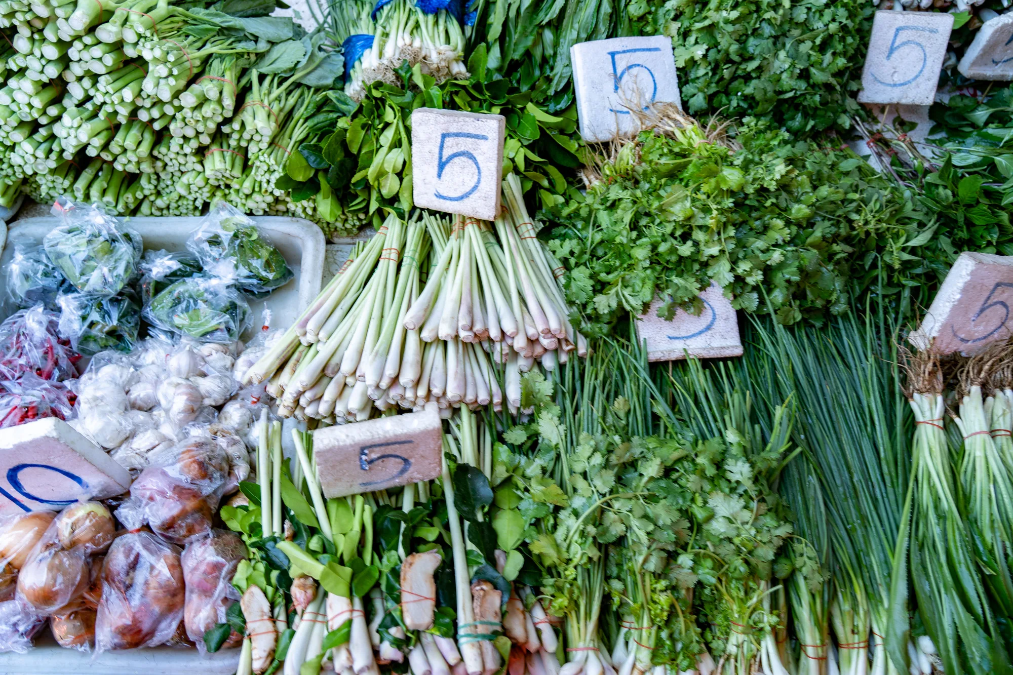 写真はタイのバンコクの市場で撮影されたものです。画面いっぱいに、様々な種類の新鮮な野菜が並べられています。左上には、緑色の葉物野菜が束ねられ、積み重ねられています。手前には、ビニール袋に入ったタマネギやニンニク、唐辛子などが見えます。写真中央には、レモングラスが束になって置かれており、その上に「5」と書かれた価格表示のプレートがあります。その他、パクチーやネギなど、様々なハーブ類が、緑鮮やかに並んでいます。全体的に活気があり、タイ料理に使われる食材の豊かさを感じさせる写真です。