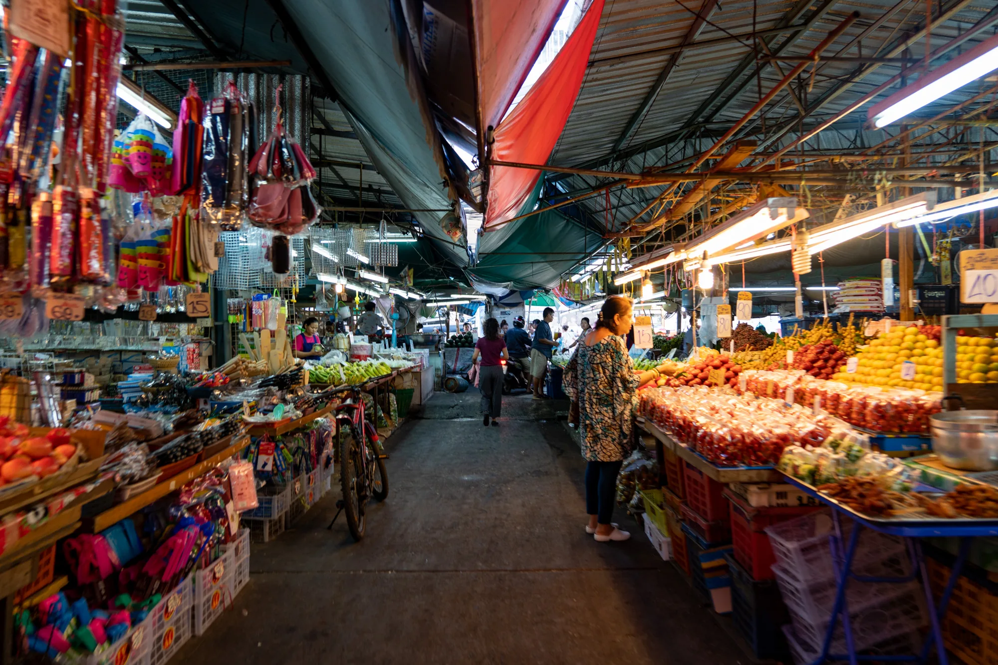 この写真はタイのバンコクで撮影された市場の様子を捉えています。市場の通路は奥へと続いており、屋根はトタンや布で覆われています。通路の両側には店が並び、色とりどりの商品が所狭しと並べられています。左側の店には、おもちゃ、文具、雑貨などが吊り下げられており、右側の店には、フルーツ、野菜、タイ料理などの食品が積み上げられています。通路を歩く人々や店員たちの姿が見え、活気ある市場の雰囲気が伝わってきます。照明は蛍光灯や電球が使われており、全体的にやや薄暗いですが、商品の鮮やかな色彩が目を引きます。