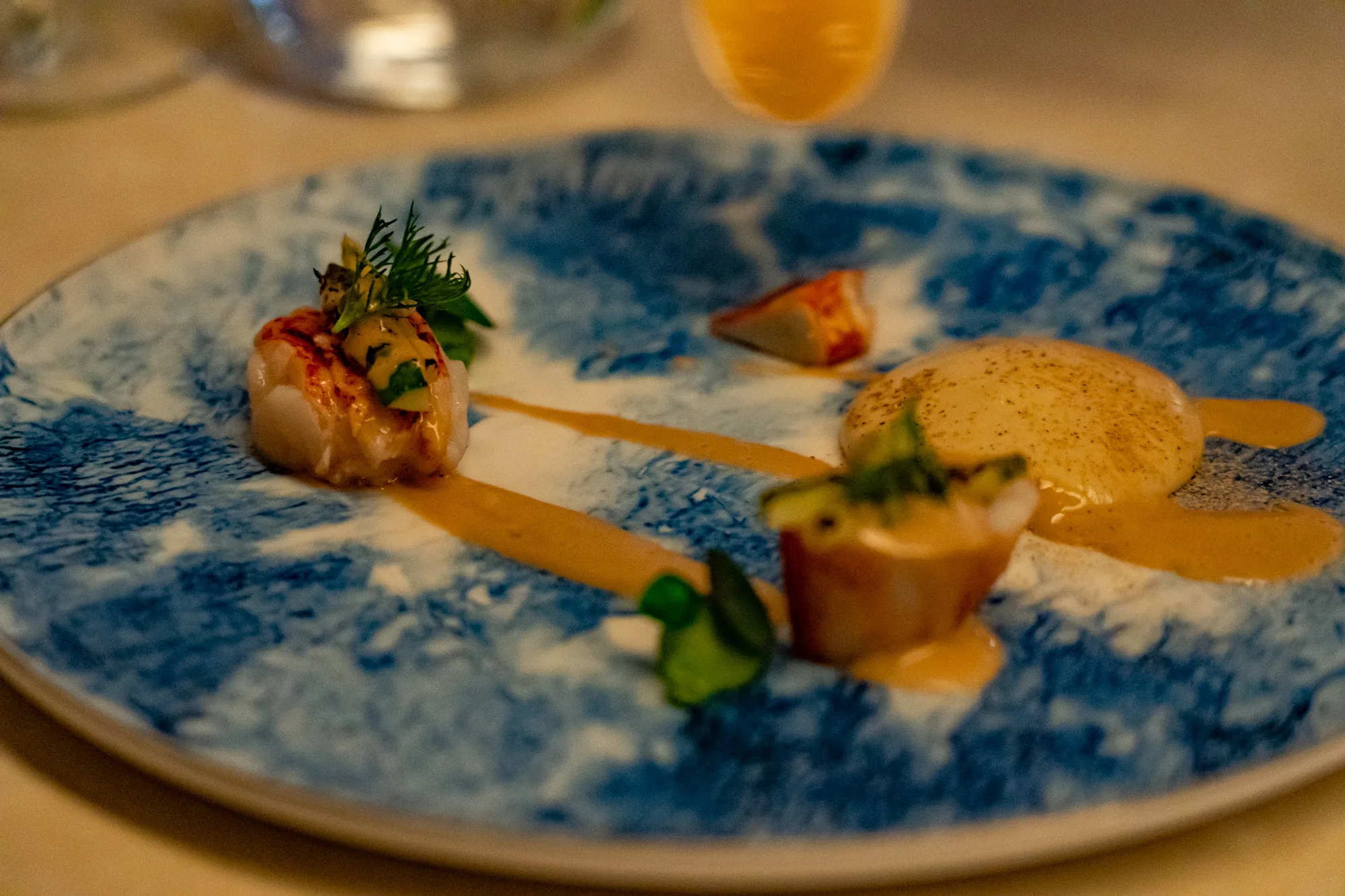 写真はタイのバンコクで撮影されたもので、高級レストランで提供されるような繊細な盛り付けの料理が写っています。料理は、青と白の模様が特徴的な皿に盛り付けられています。皿の上には、エビまたはロブスターのような魚介類を使った料理が、丁寧に配置されています。２つの魚介類のポーションには、それぞれハーブや野菜が添えられ、美しい彩りを添えています。さらに、滑らかなソースが皿全体に流れ、料理の風味を引き立てているようです。皿の中央には、丸いムースのようなものが置かれ、軽くスパイスが振られています。全体的に、上品で洗練された雰囲気が漂っており、特別な日に味わいたい高級料理であることが伺えます。