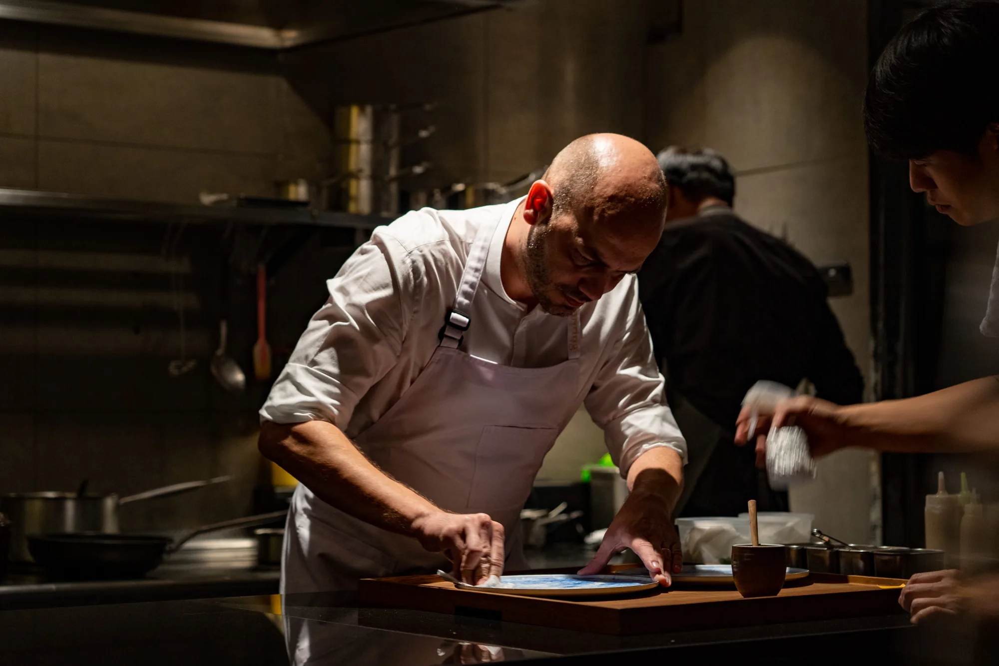 写真はタイのバンコクで撮影された厨房の様子を捉えています。中央にいるのは、白いコックコートとエプロンを身につけたシェフで、テーブルの上に置かれた青い模様の皿を丁寧に磨いているようです。シェフは短髪で、ひげを生やしており、真剣な表情で作業に取り組んでいます。  背景には、ステンレス製の調理器具や棚が見え、厨房の機能的な雰囲気を醸し出しています。シェフの奥には、別のコックコートを着た人物のシルエットがあり、複数人が忙しく立ち働いている様子が伺えます。手前には、料理の準備に必要なと思われる小さなカップや調味料などが置かれています。暗めの照明が、シェフの作業に集中する様子を強調しており、プロフェッショナルの緊張感と調理への情熱を感じさせる一枚です。