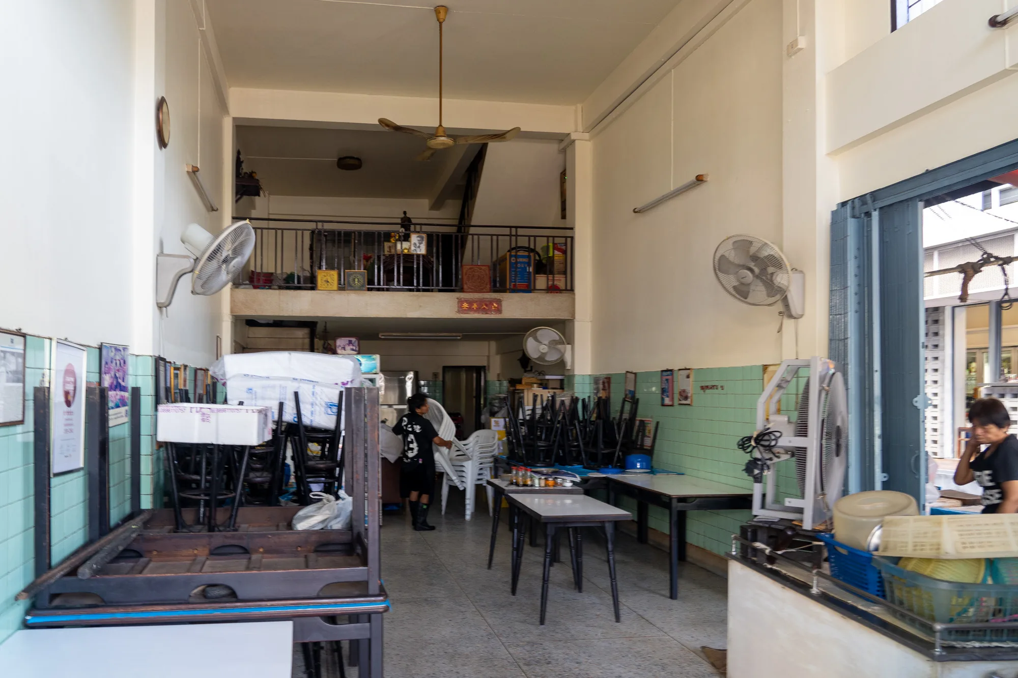 写真はタイのバンコクで撮影されたと思われる、ローカルな食堂の内部を捉えています。全体的に昼間の自然光が入っており、明るく、どこか懐かしい雰囲気が漂っています。店内は、テーブルや椅子が積み重ねられており、営業前の準備中か、もしくは閉店後の片付け中であるように見えます。天井にはレトロなシーリングファンが設置され、壁には年季の入った扇風機や、写真、装飾品などが飾られています。二階部分には手すりが設けられており、雑貨などが置かれているのが見えます。店の入り口付近には人が立っており、外の様子を伺っているようです。床はタイル張りで、年季が入っていることがわかります。全体的に少し雑然とした雰囲気ですが、タイのローカルな日常が垣間見える写真です。