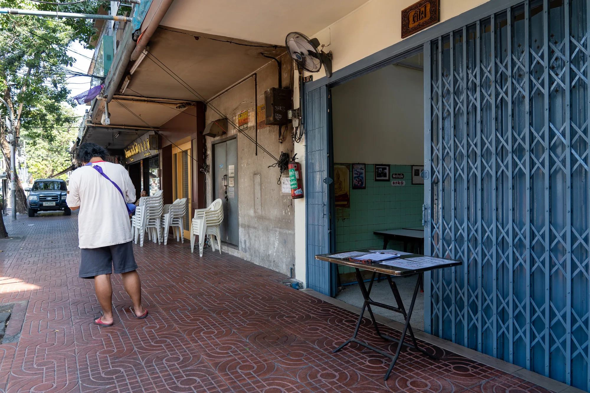 写真はタイのバンコクで撮影された、街中の日常的な風景を捉えています。画面左側には、木が並ぶ道路と一台の自動車が写っています。歩道は赤茶色のタイルで舗装されており、独特の模様が施されています。  歩道沿いには、建物が並んでおり、その一部は商店や飲食店であるようです。手前には、白いシャツと灰色の短パンを身に着けた男性が歩いており、紫色のバッグを肩にかけています。建物の軒下には、白いプラスチック製の椅子が積み重ねられています。  右側には、シャッターが開いたお店の入り口が見えます。青いシャッターは一部開かれており、店内の様子がわずかに伺えます。入り口の脇には、折り畳み式のテーブルが置かれ、書類のようなものが置かれています。店内は緑色のタイルで装飾されており、壁には写真や額縁が飾られています。壁には扇風機が取り付けられています。  全体的に、写真はタイのバンコクにおける日常の生活風景を捉えており、ローカルな雰囲気が感じられます。