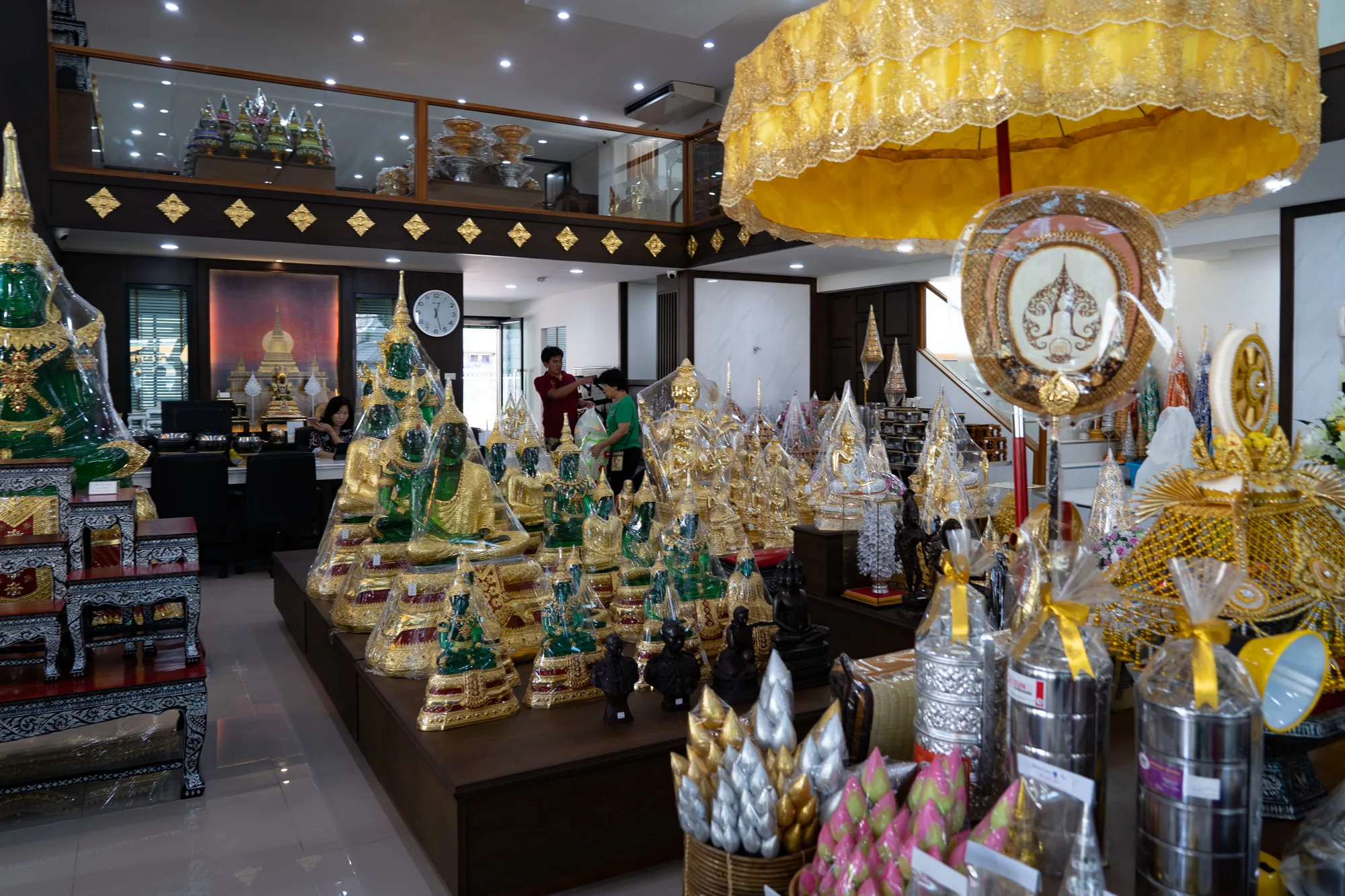 写真はタイのバンコクで撮影されたもので、仏像や仏教関連の装飾品を扱うお店の内部を捉えています。店内には、金、緑、黒など様々な色合いの仏像が多数並べられています。多くはビニールで包装されており、新品であることがわかります。仏像の他にも、黄金色の傘のような装飾品や、蓮の花を模した飾り、銀色の弁当箱のようなものが陳列されています。お店の奥にはレジらしき場所があり、店員らしき人物が数名見えます。店内は明るく、天井には照明が設置されています。落ち着いた色調の木製の棚や内装が、商品である仏像の鮮やかさを引き立てています。上階にはガラス張りのスペースがあり、そこにも様々な装飾品が飾られているのが見えます。全体的に、タイの伝統的な宗教文化を感じさせる、きらびやかで華やかな雰囲気の写真です。