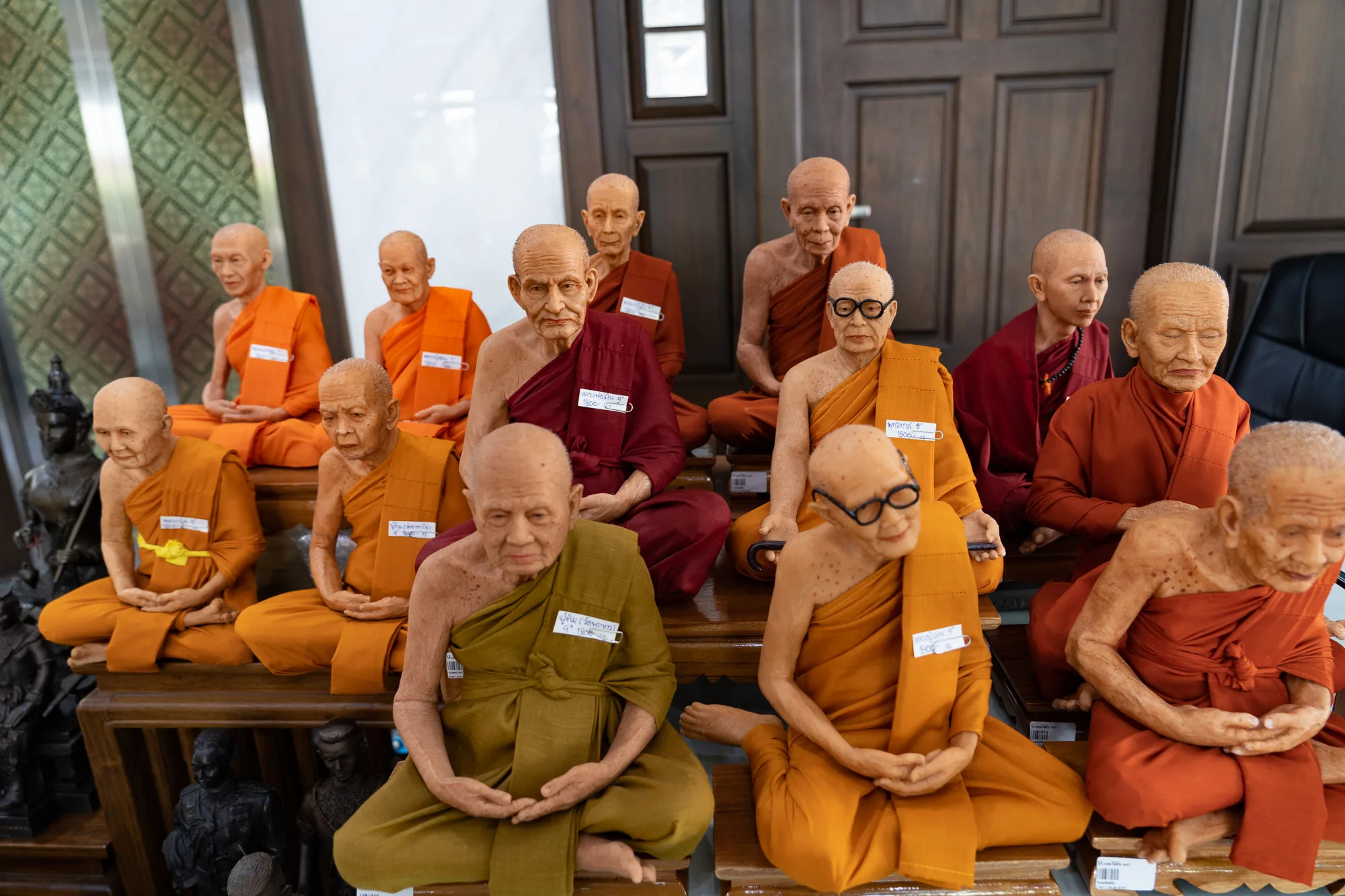 タイのバンコクで撮影された写真です。写真には、様々な色の僧衣をまとった僧侶の人形が複数並んでいます。人形は写実的に作られており、それぞれ表情や姿勢が異なっています。僧侶の人形は、木製の台座の上に座っており、背景には扉のようなものが写っています。人形には名前や数字が書かれた札がつけられています。落ち着いた雰囲気で、宗教的な空間を彷彿とさせます。