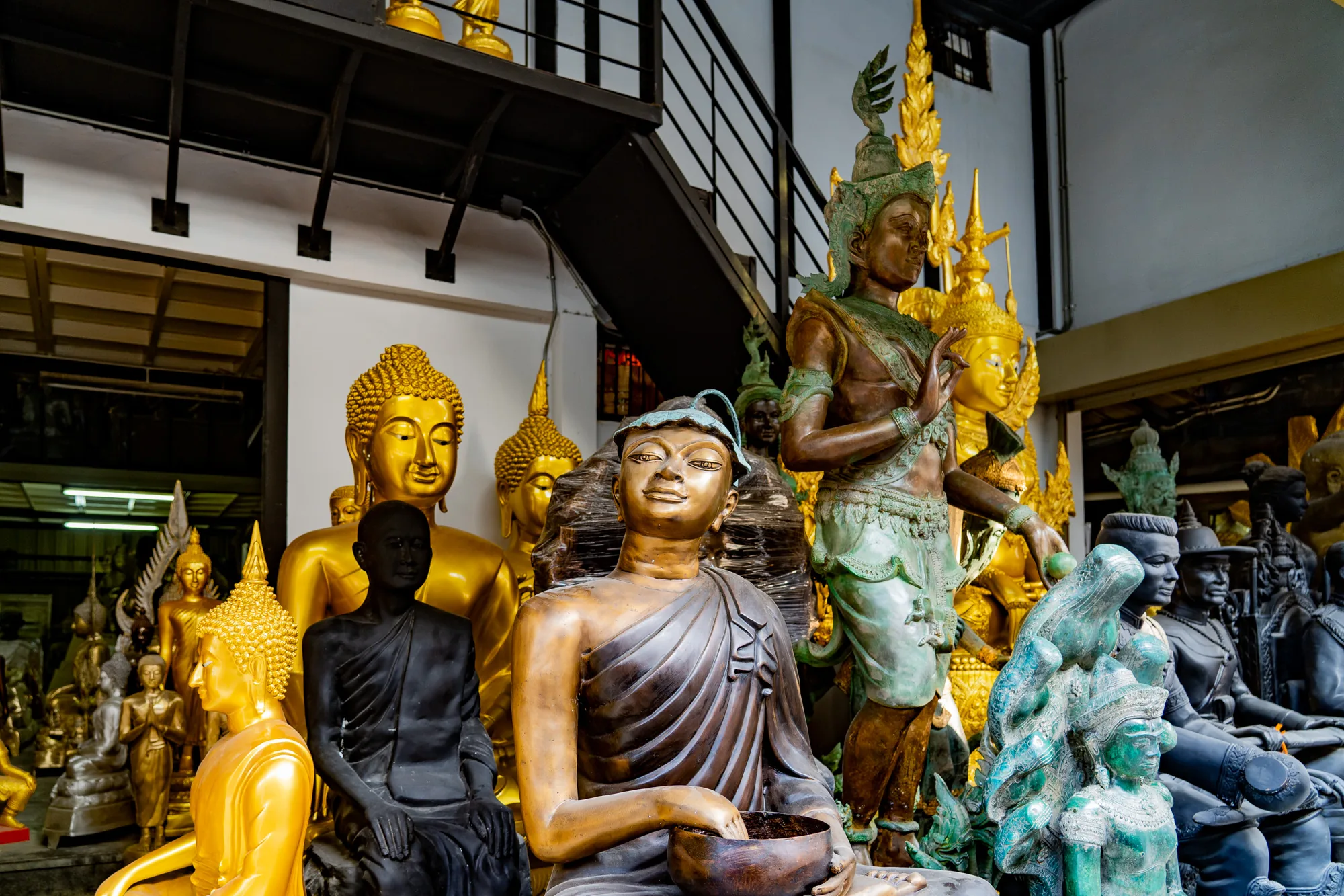 写真はタイのバンコクで撮影されたもので、店内には多様な仏像や像が並んでいます。これらの像は金色、ブロンズ色、黒色、緑色など様々な色調を持ち、仏陀の像、僧侶の像、神像など、表現されている存在も様々です。  背景には建物の内部構造が見え、階段や金属製の梁が確認できます。これは、写真が屋内、おそらく仏像を販売する店内で撮影されたことを示唆しています。  写真全体の雰囲気は、神聖で静謐であり、タイの文化や宗教的な要素が強く感じられます。光の当たり具合や像の配置が、その雰囲気をさらに強調しています。