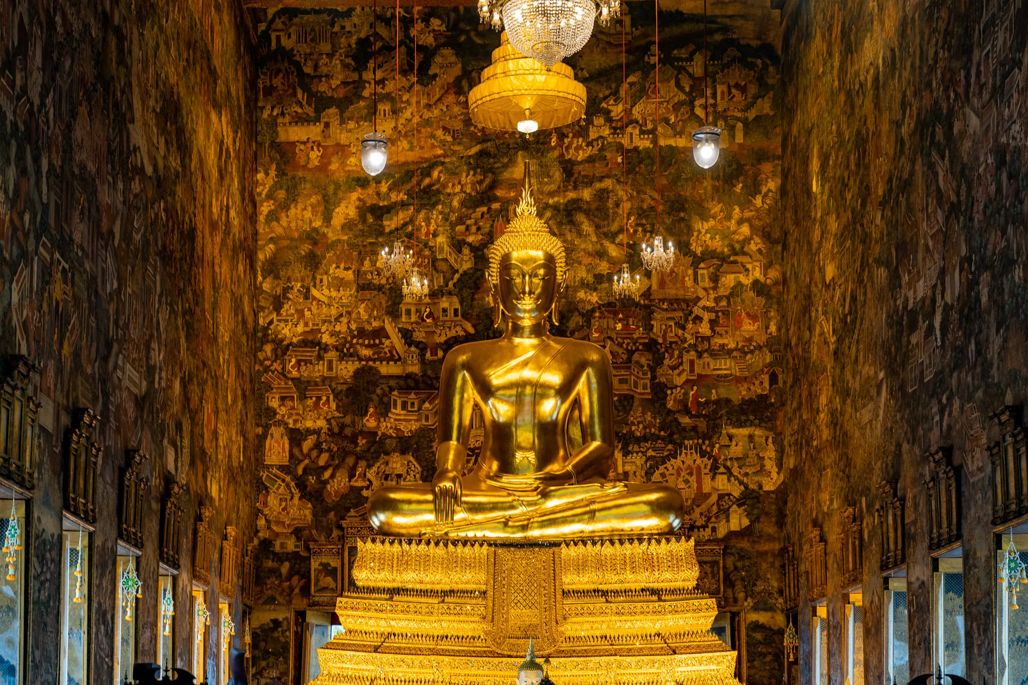写真はタイのバンコクにある寺院の内部を捉えたものです。中央には、大きく輝く黄金の仏像が鎮座しています。仏像は光沢があり、堂々とした印象を与えます。仏像の背後には、壁一面に細密な壁画が描かれています。壁画は金色を基調としており、古代の生活や神話を表現しているようです。天井からはシャンデリアが吊り下げられ、空間全体を明るく照らしています。窓からは自然光が差し込み、厳かな雰囲気を醸し出しています。全体的に、豪華絢爛で神聖な空間であることが伝わってきます。