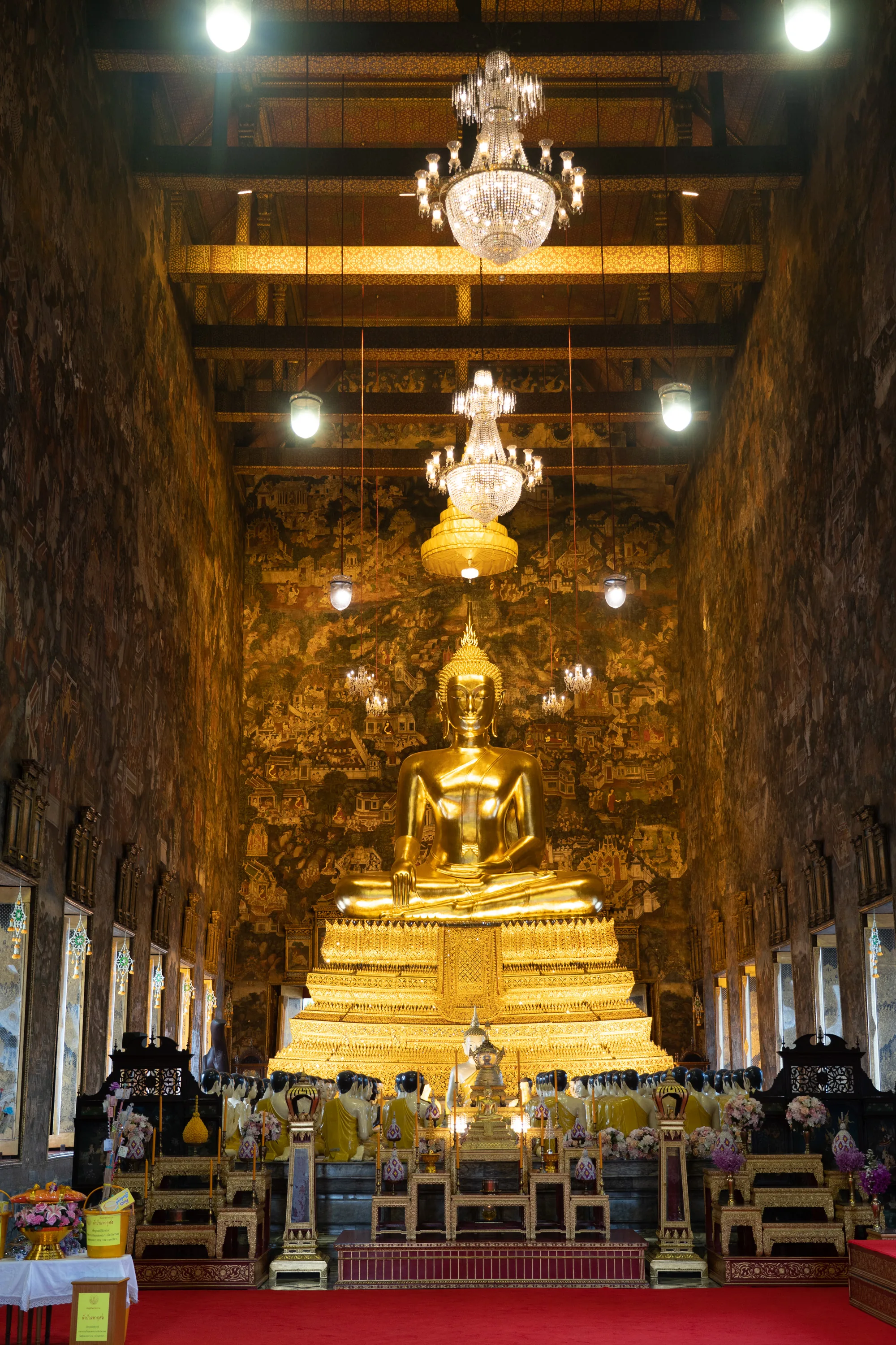 写真はタイのバンコクにある寺院内部を写しています。中央には巨大な黄金の仏像が鎮座しており、圧倒的な存在感を放っています。仏像は台座の上に安置され、さらにその手前には供物が捧げられています。  壁面には歴史的な出来事を描いた壁画が描かれており、天井からは豪華なシャンデリアが吊り下げられています。シャンデリアの光が仏像を照らし出し、神聖な雰囲気を醸し出しています。  写真全体は金色を基調としており、豪華絢爛でありながらも、静寂で落ち着いた雰囲気が感じられます。窓から差し込む光が空間に奥行きを与え、荘厳さを際立たせています。