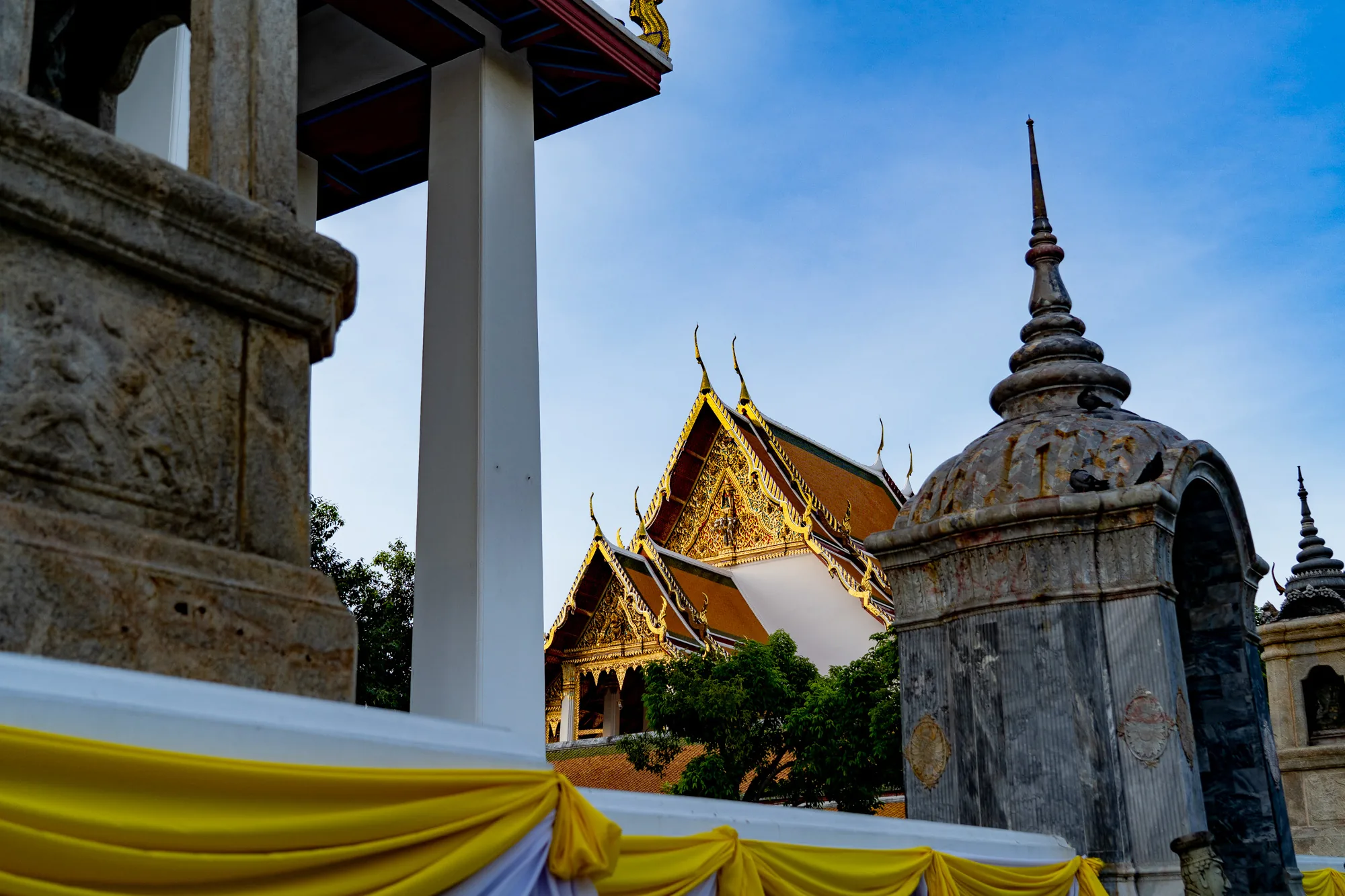 写真はタイのバンコクで撮影されたものと思われます。青い空を背景に、伝統的なタイ寺院の建築様式が捉えられています。前景には、石造りの装飾的な建造物の一部がぼやけて写っており、黄色の布で飾られています。中央には、鮮やかな金色とオレンジ色の装飾が施された屋根が特徴的な寺院が見えます。その建築様式は、タイの寺院独特の尖った屋根と細部にわたる装飾を反映しています。寺院の周りには緑豊かな木々が生い茂っており、自然と調和した寺院の様子が伺えます。右側には、石造りの仏塔のような構造物があり、歴史を感じさせる風合いです。写真全体を通して、タイの文化的な豊かさと美しい建築様式が表現されています。