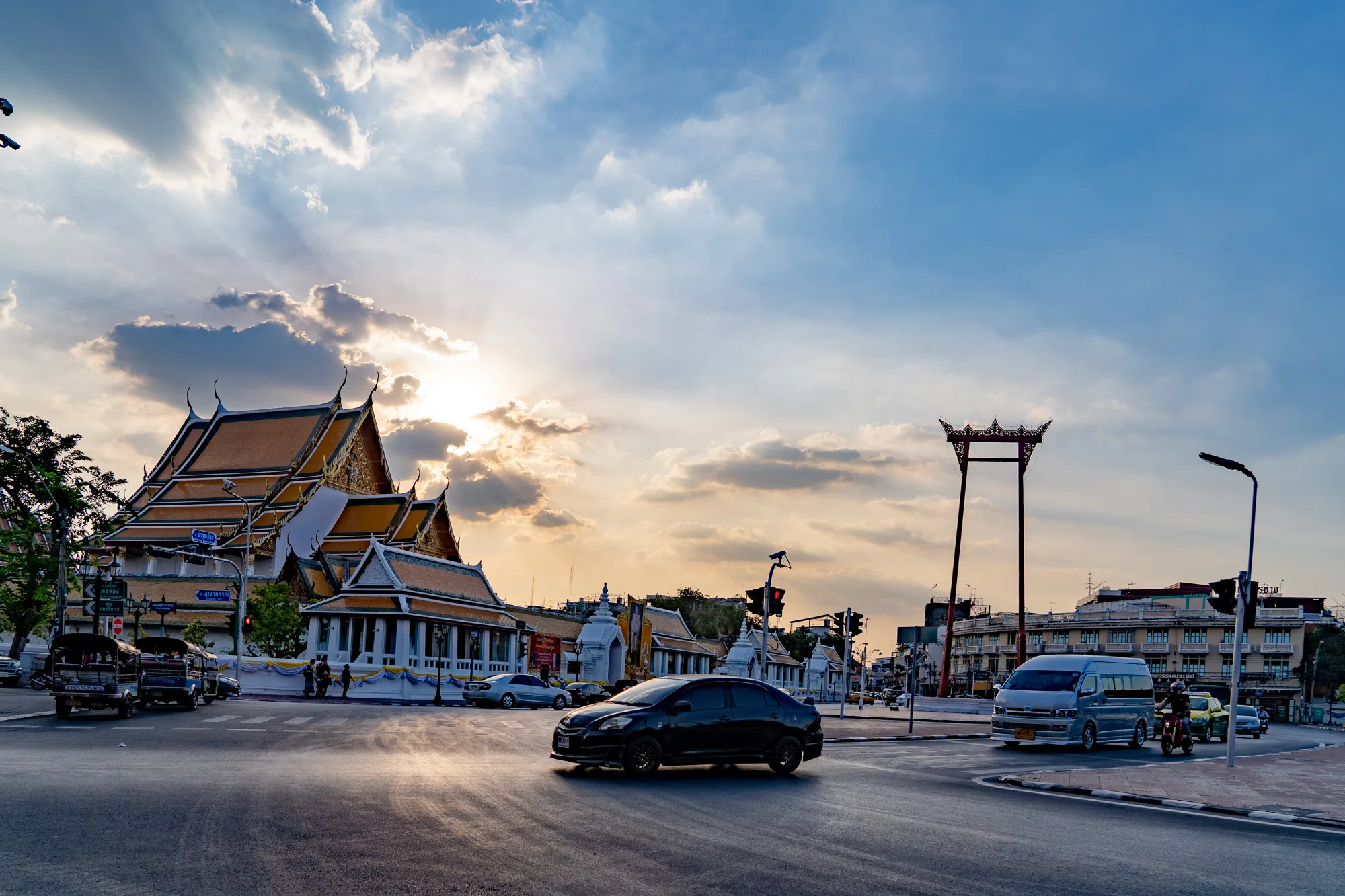 写真はタイのバンコクで撮影されたもので、夕暮れ時の街並みが写っています。空には雲が広がり、雲間から太陽光が差し込んでいます。写真の中央には、特徴的な屋根を持つタイの伝統的な寺院が見えます。寺院の周りには白い壁があり、内部を伺うことはできません。寺院の隣には、象徴的な巨大ブランコである「Giant Swing（ブランコ）」がそびえ立っています。  道路には、黒い車や銀色のバン、トゥクトゥクなど、様々な種類の車が走っています。また、信号機や街灯、標識なども確認できます。写真全体は、夕暮れ時の穏やかな光に包まれており、バンコクの活気と伝統が調和した雰囲気を醸し出しています。交通量は比較的多いですが、騒がしさを感じるような雰囲気ではありません。全体的に落ち着いた印象の写真です。