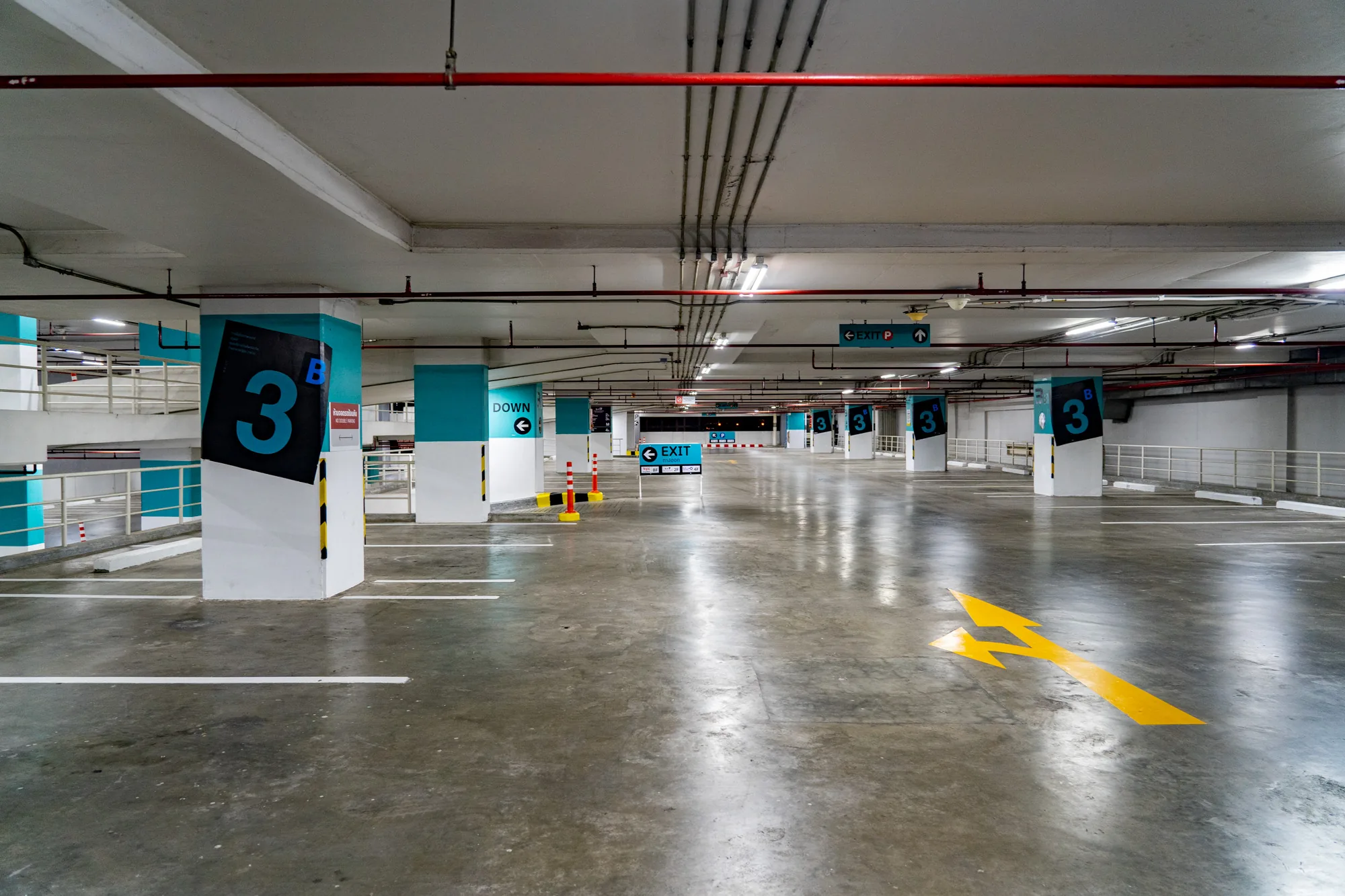 写真はタイのバンコクで撮影された、屋内駐車場の様子を写しています。駐車場はがらんとしており、ほとんど車が停まっていません。床は磨かれたコンクリートで、光を反射しています。駐車スペースを示す白い線が引かれており、床には出口の方向を示す黄色い矢印が描かれています。柱は白く塗られ、ターコイズブルーのアクセントがあり、黒地に水色の数字「3」が記された標識が取り付けられています。天井は白く、赤色の配管が走っています。また、「EXIT」や駐車場への案内を示す標識も見られます。全体的に、清潔で整理された印象の駐車場です。