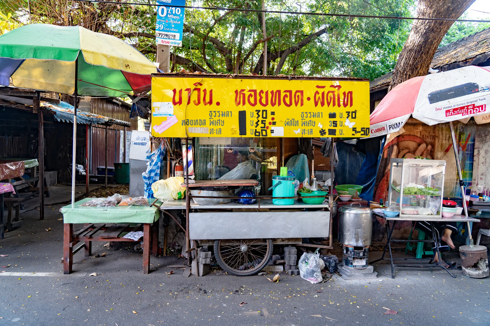 この写真はタイのバンコクで撮影されたものと思われます。道路に面した屋台が写っており、アジアらしい活気と少しの雑多さを感じさせます。屋台には大きな黄色い看板があり、タイ語でメニューと価格が書かれています。「หอยทอด ผัติไท（ホイトート パッタイ）」と読める部分があるので、牡蠣の鉄板焼き（ホイトート）やパッタイなどを提供している屋台でしょう。  屋台は簡素な作りで、テーブルや椅子、調理器具などが置かれています。周囲にはパラソルが立てられており、日差しを遮っています。左側のテーブルには、何か食品が並べられているのが見えます。右側のテーブルには、ガラスケースに入った食材が見えます。屋台の奥には人が座っているのが見え、調理をしているのかもしれません。路上には落ち葉が散らばっており、生活感のある風景です。